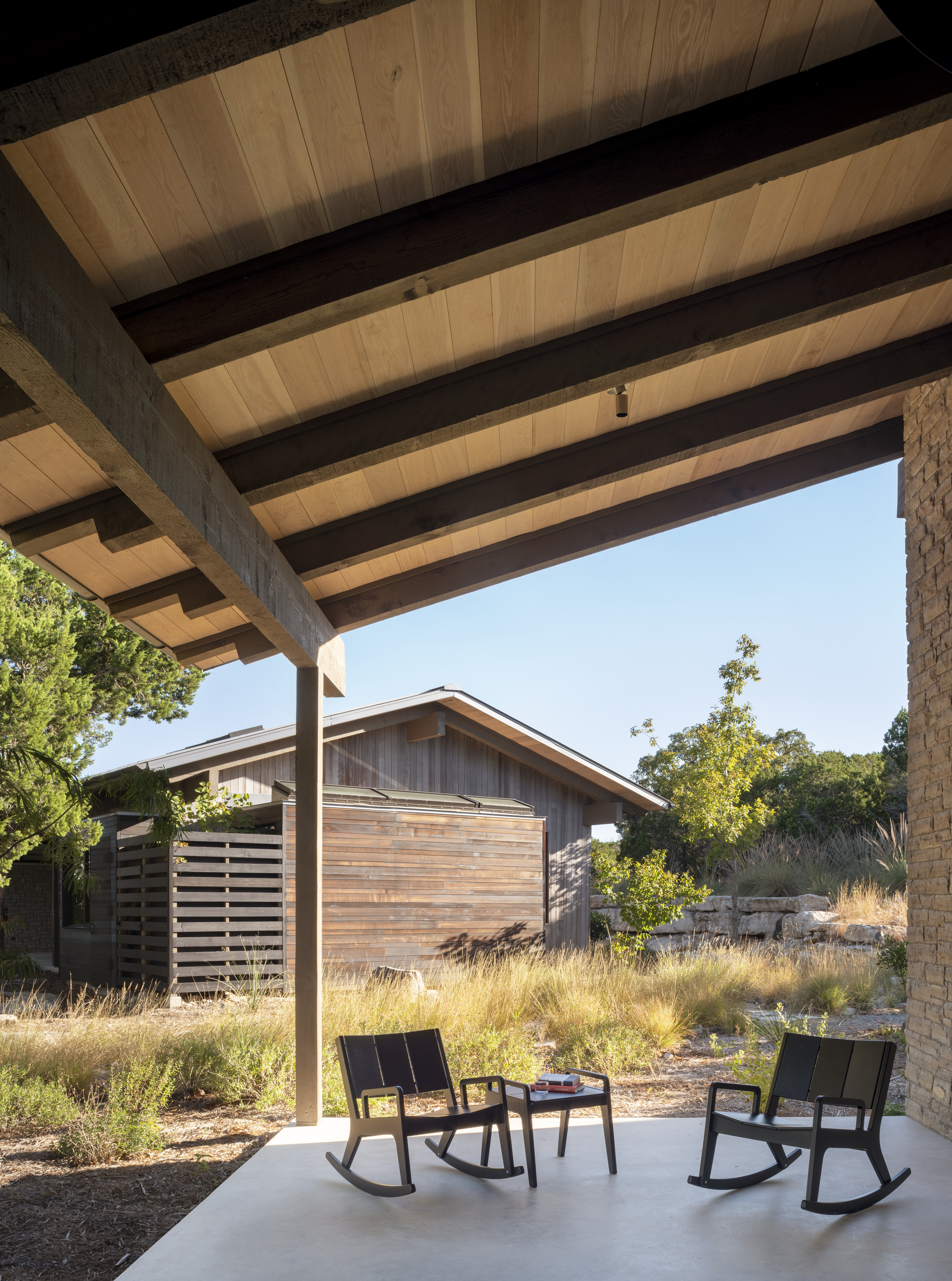 A porch with a wooden roof and four black rocking chairs overlooks a garden with wild grasses and a wooden shed in the background, reminiscent of the serene atmosphere at Laity Lodge's Cedar Brake.