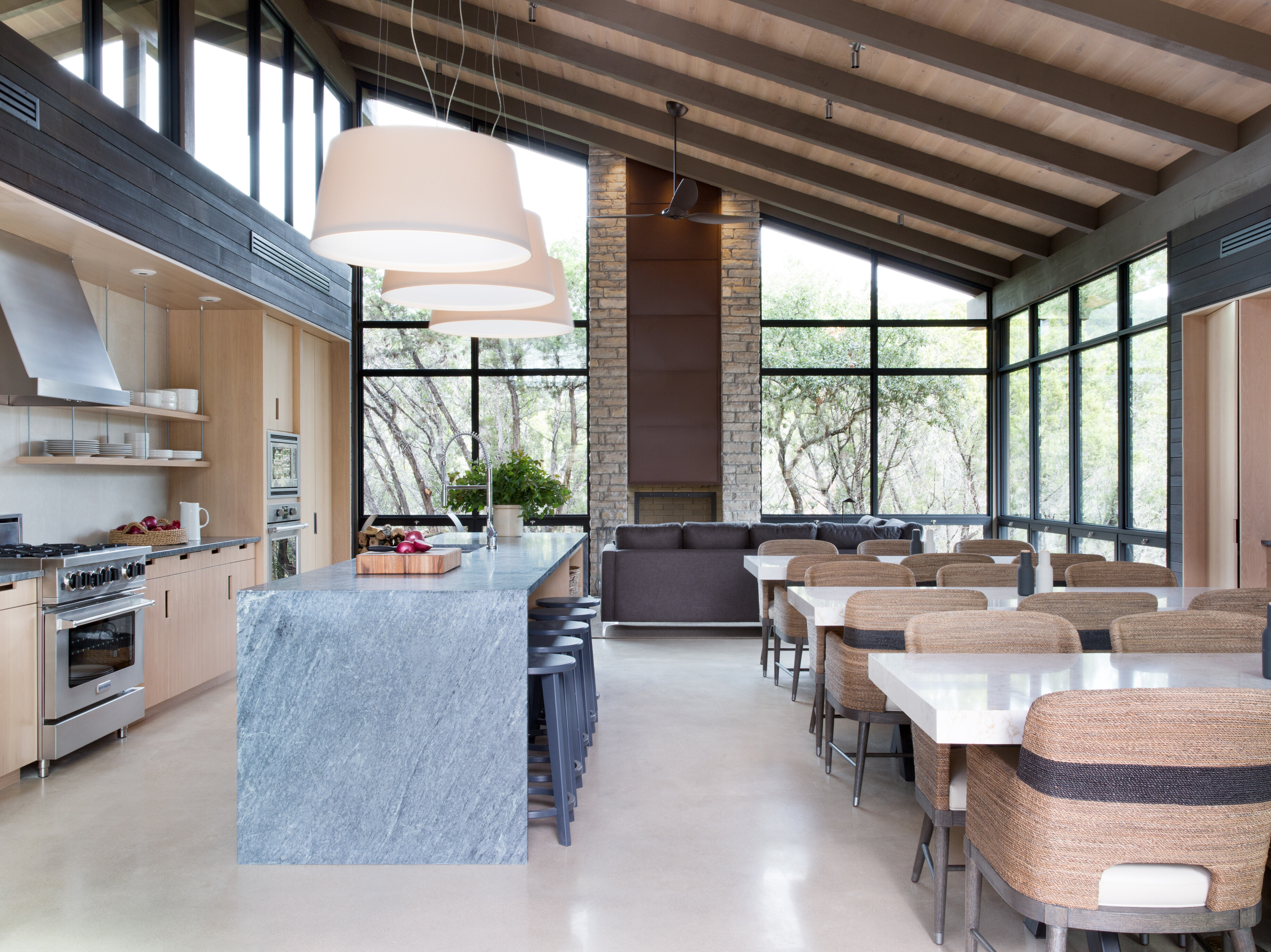 Spacious modern kitchen and dining area with large windows, an island with barstools, a stone countertop, contemporary furniture, and a high angled ceiling with exposed beams reminiscent of Cedar Brake's rustic charm at Laity Lodge.