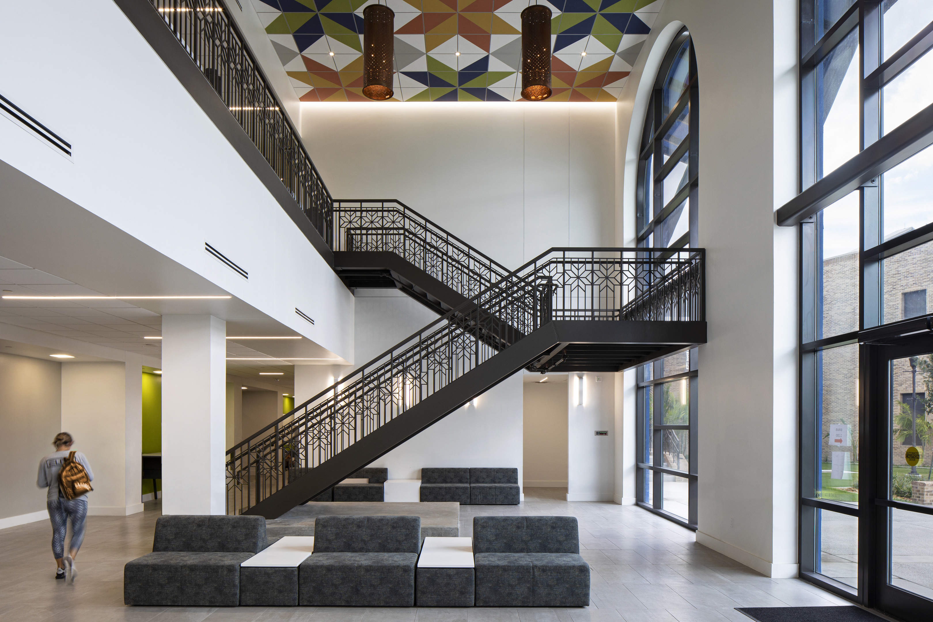 A person walks in a spacious, modern atrium featuring a geometric ceiling design, large windows, a staircase with patterned railings, and grey, modular seating.