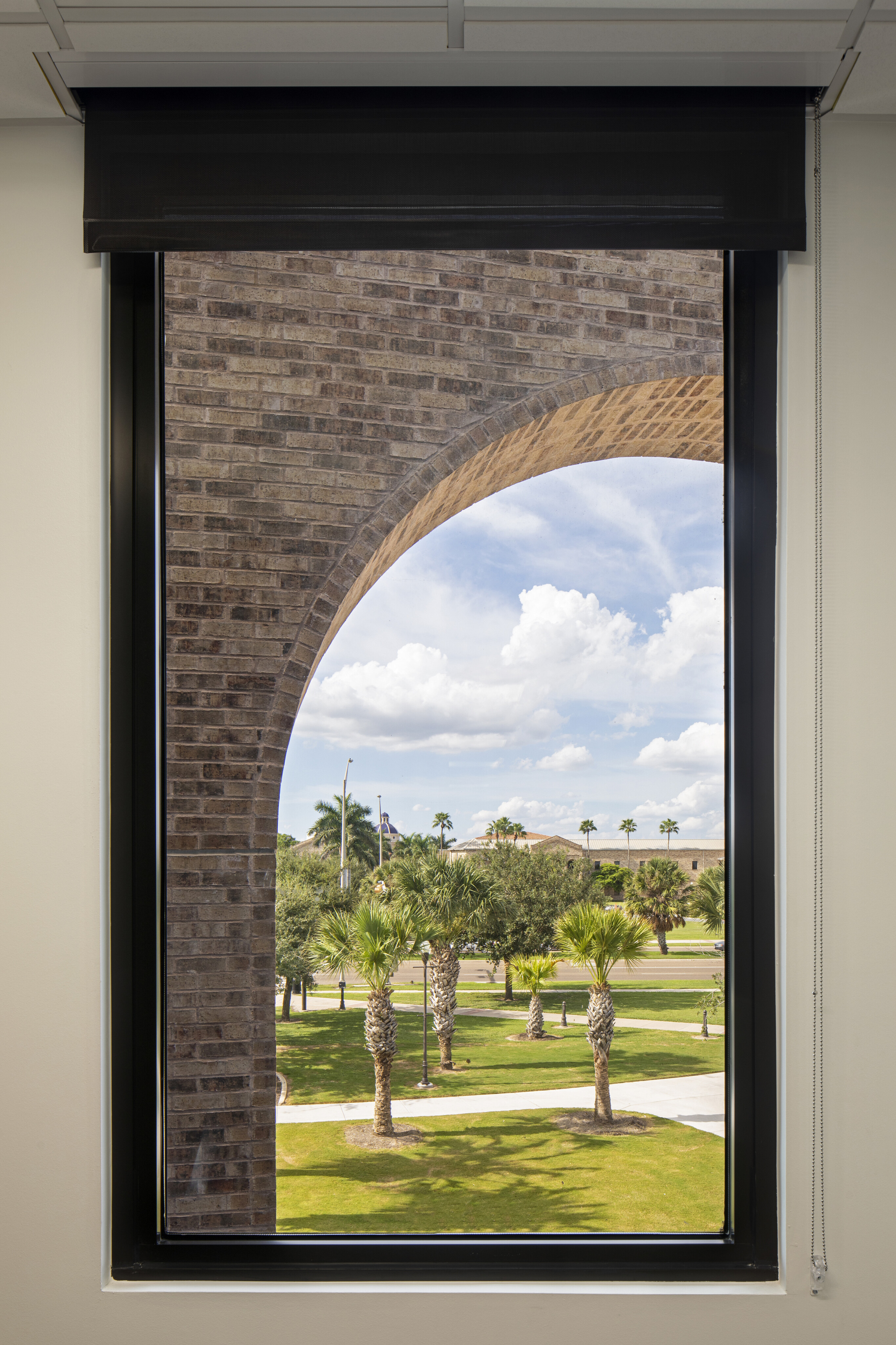 View through a rectangular window with a raised blind, showing an outdoor scene with a brick arch, palm trees, green grass, and a partly cloudy sky.