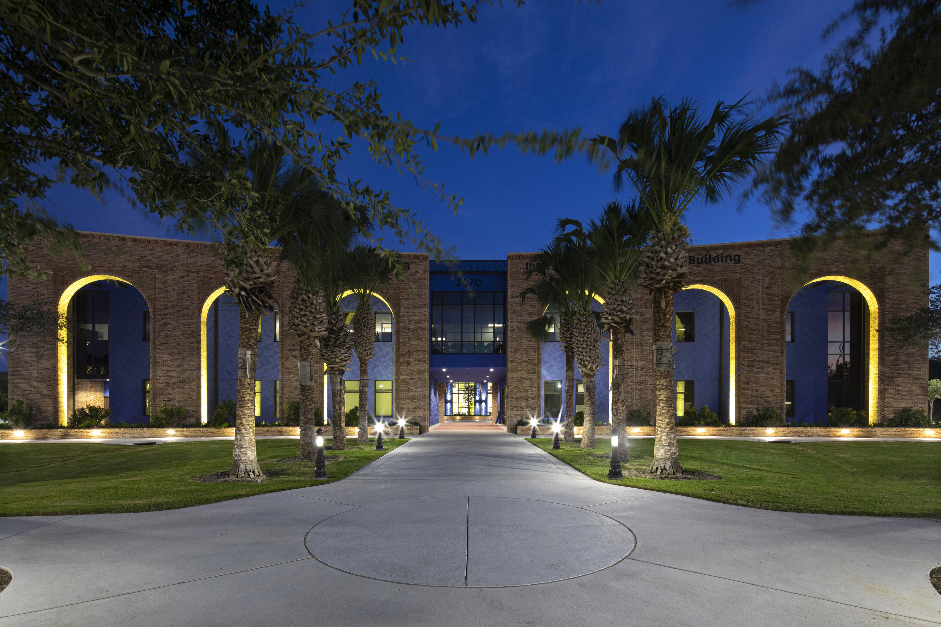 A modern building with a curved facade, illuminated at night. Pathway and palm trees lead to the entrance of the UTRGV Interdisciplinary Academic Building in Brownsville.