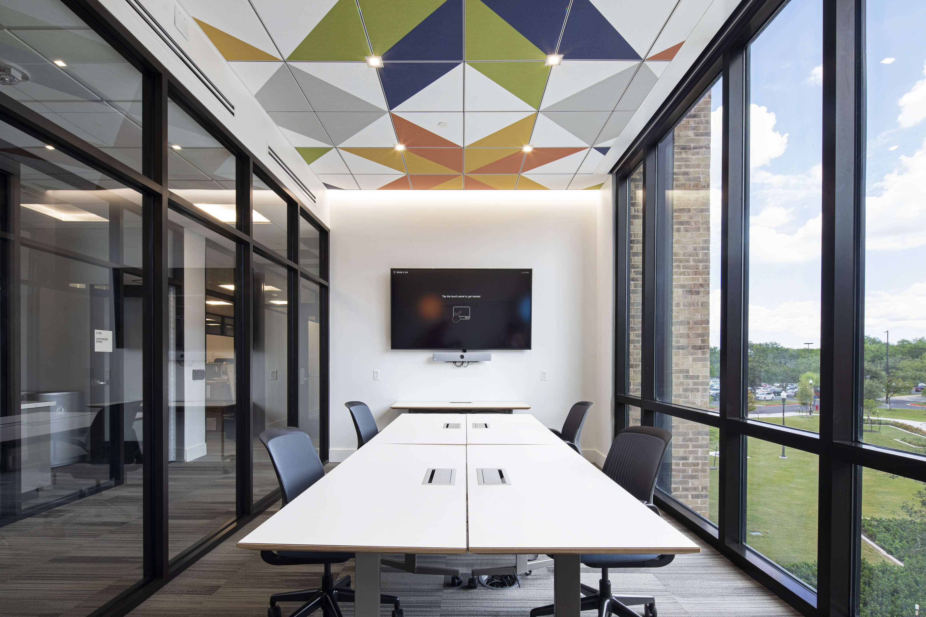 A modern conference room with a geometric ceiling design, glass walls, a wall-mounted TV, white tables, and office chairs. The room has large windows providing a view of greenery outside.