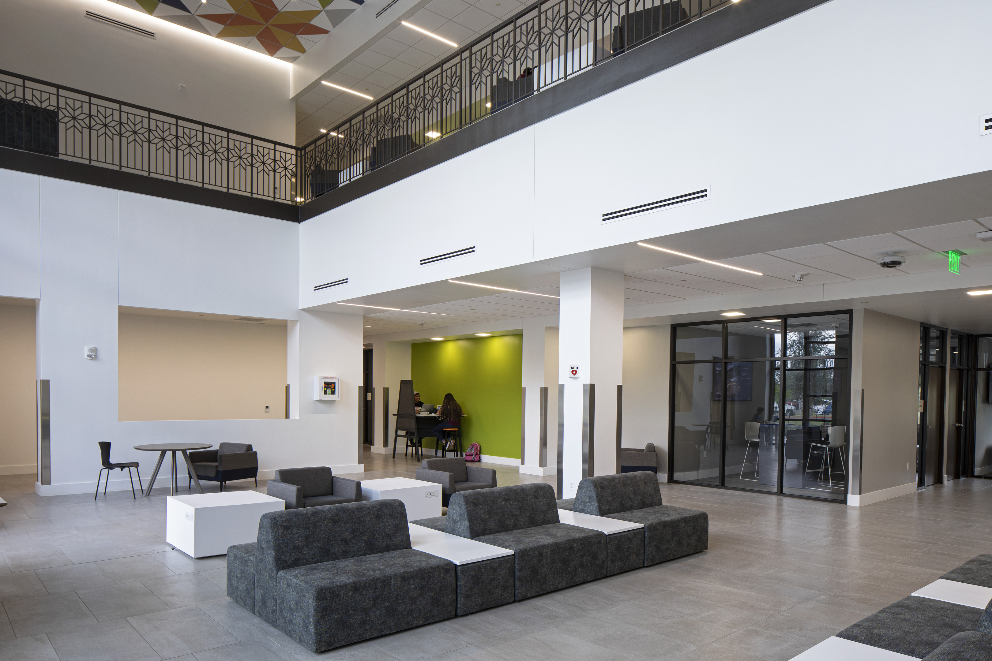 A modern and spacious two-story lobby in the UTRGV Interdisciplinary Academic Building in Brownsville features gray and white modular seating, a green accent wall, and a glass-walled room. An individual sits at a piano in the far corner near a black railing on the upper level.