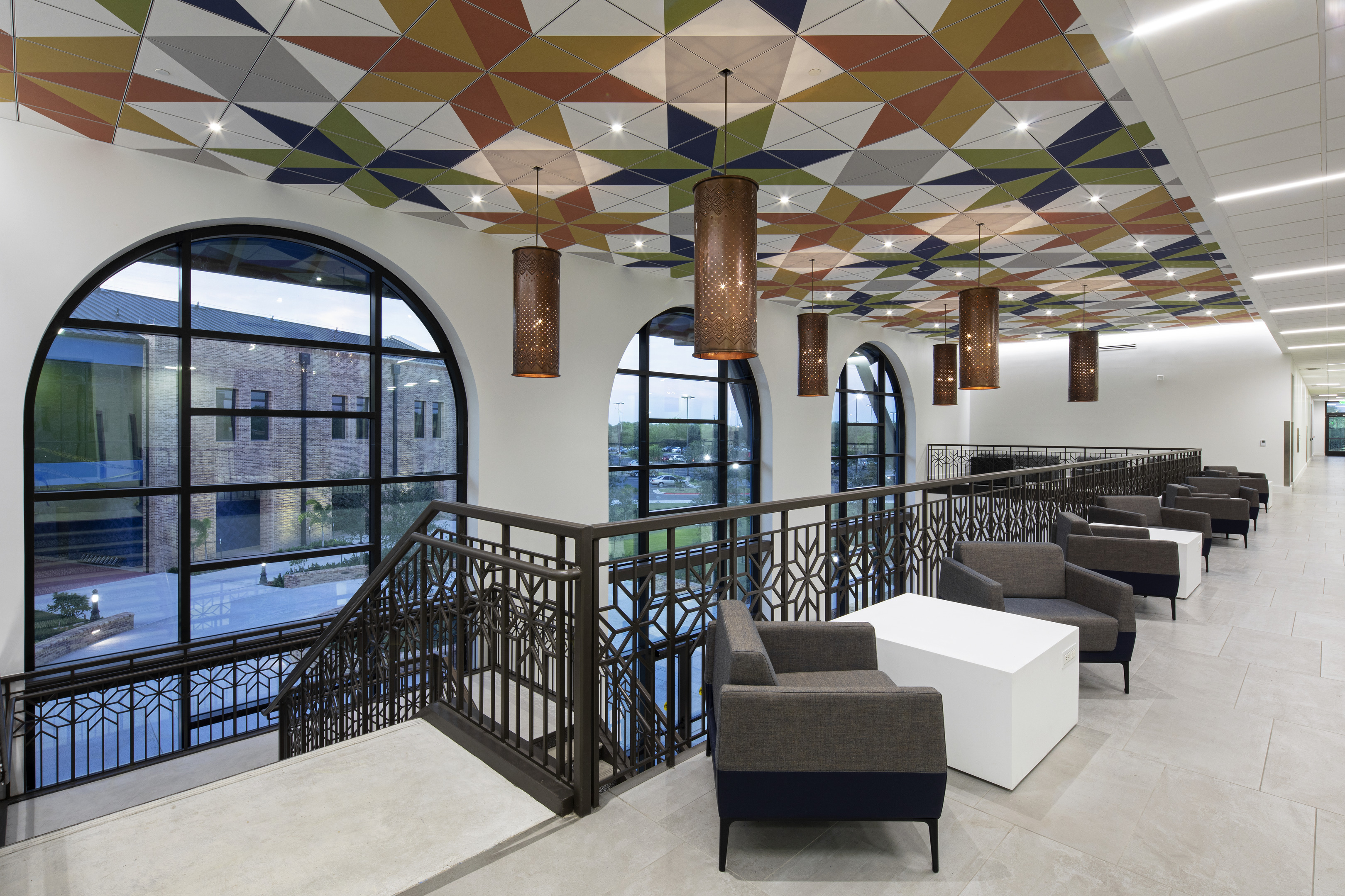 Modern indoor balcony with patterned ceiling, arched windows, and contemporary furniture including dark chairs and white tables. The view through the windows shows a building and an outdoor area.