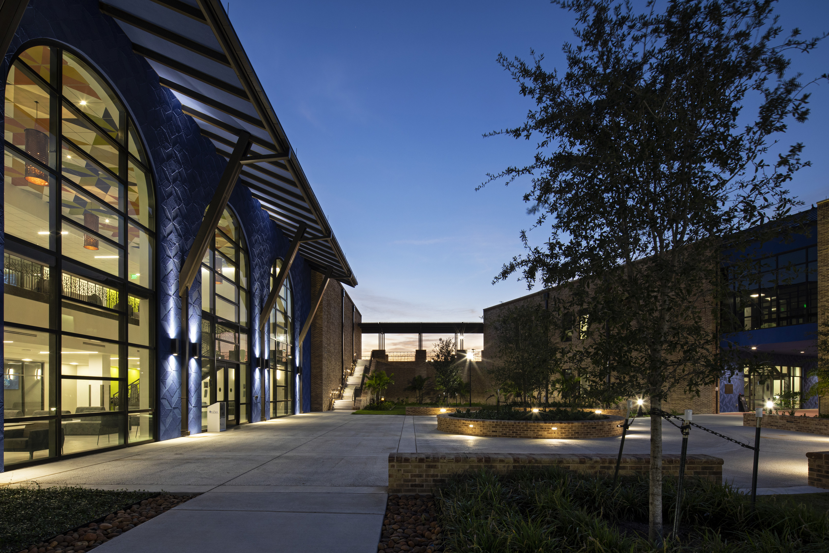 A modern building with large glass windows and blue exterior walls is illuminated at dusk. The scene includes a courtyard with trees and planters, and a lit pathway leading up to another structure.