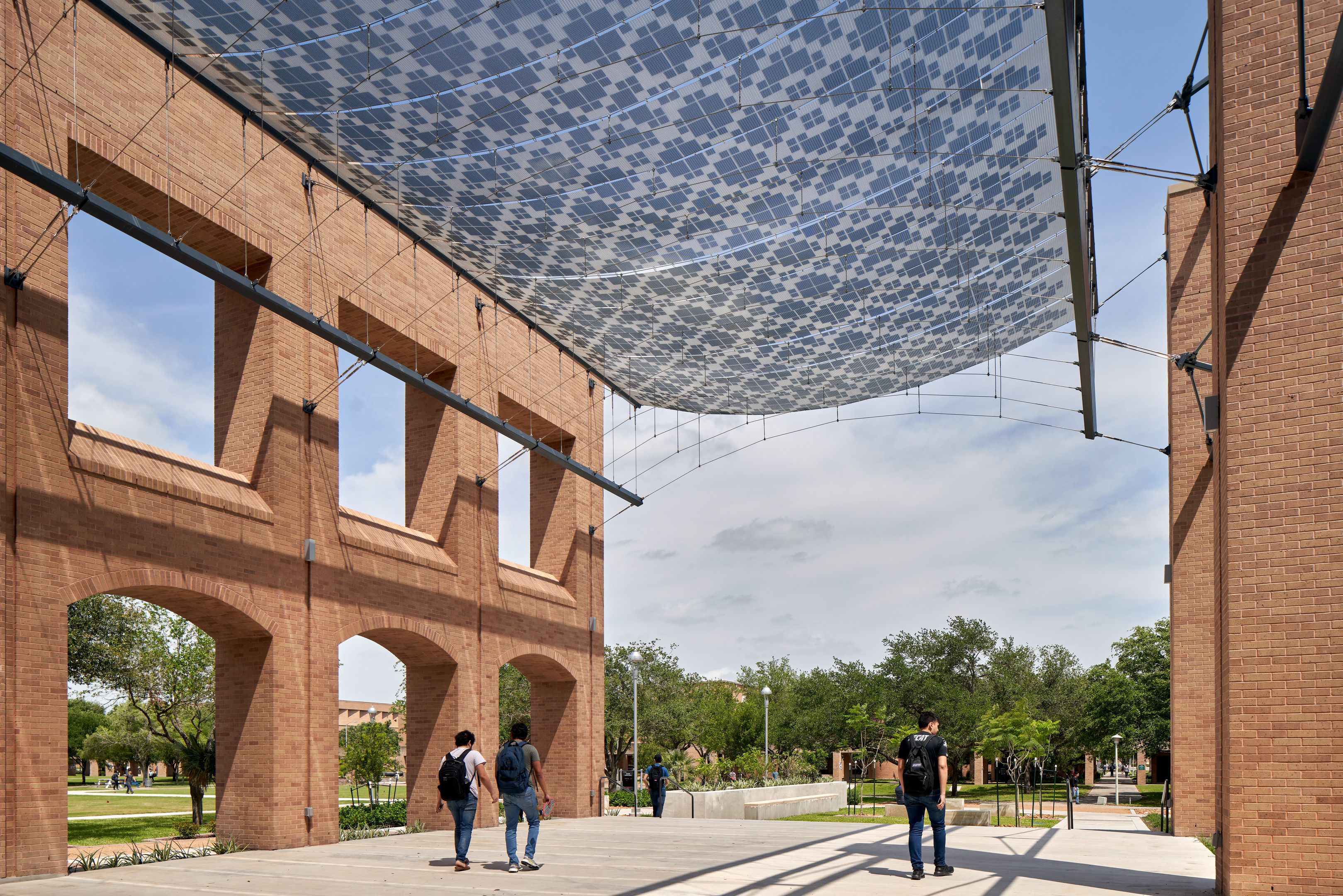 People walk under a large, mesh-like canopy structure with pixelated patterns between brick buildings on a sunny day.