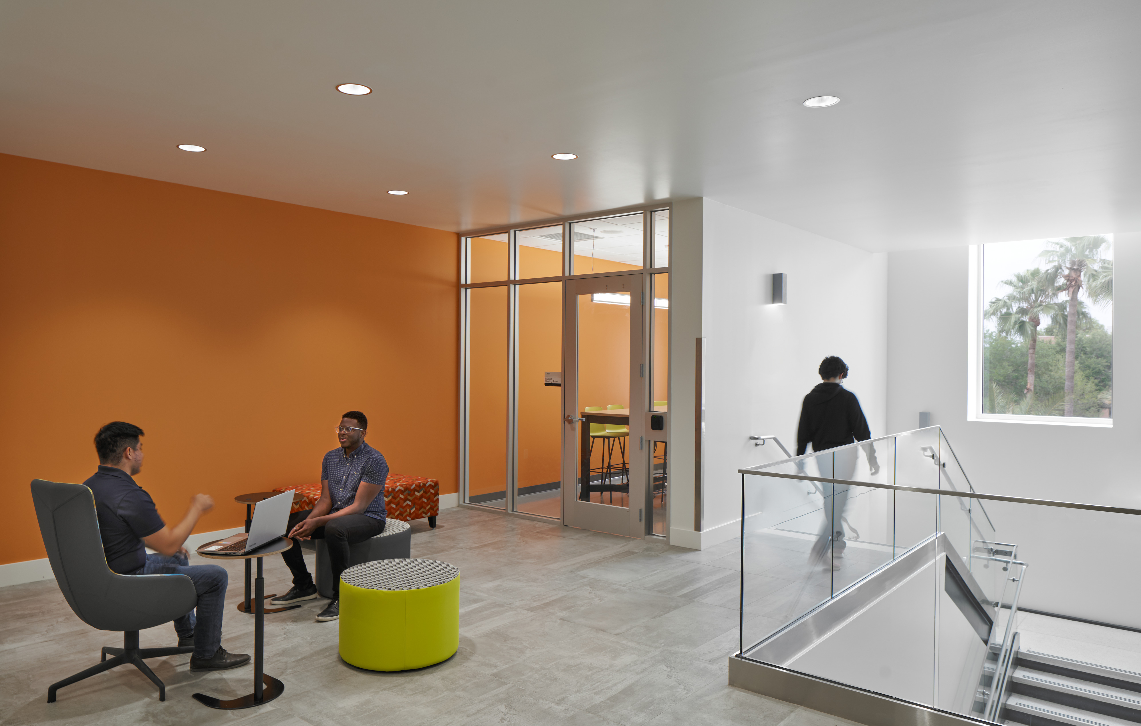 Two individuals are seated and conversing near a small table in a modern office space with orange and white walls. A person is walking down a hallway with a glass railing.