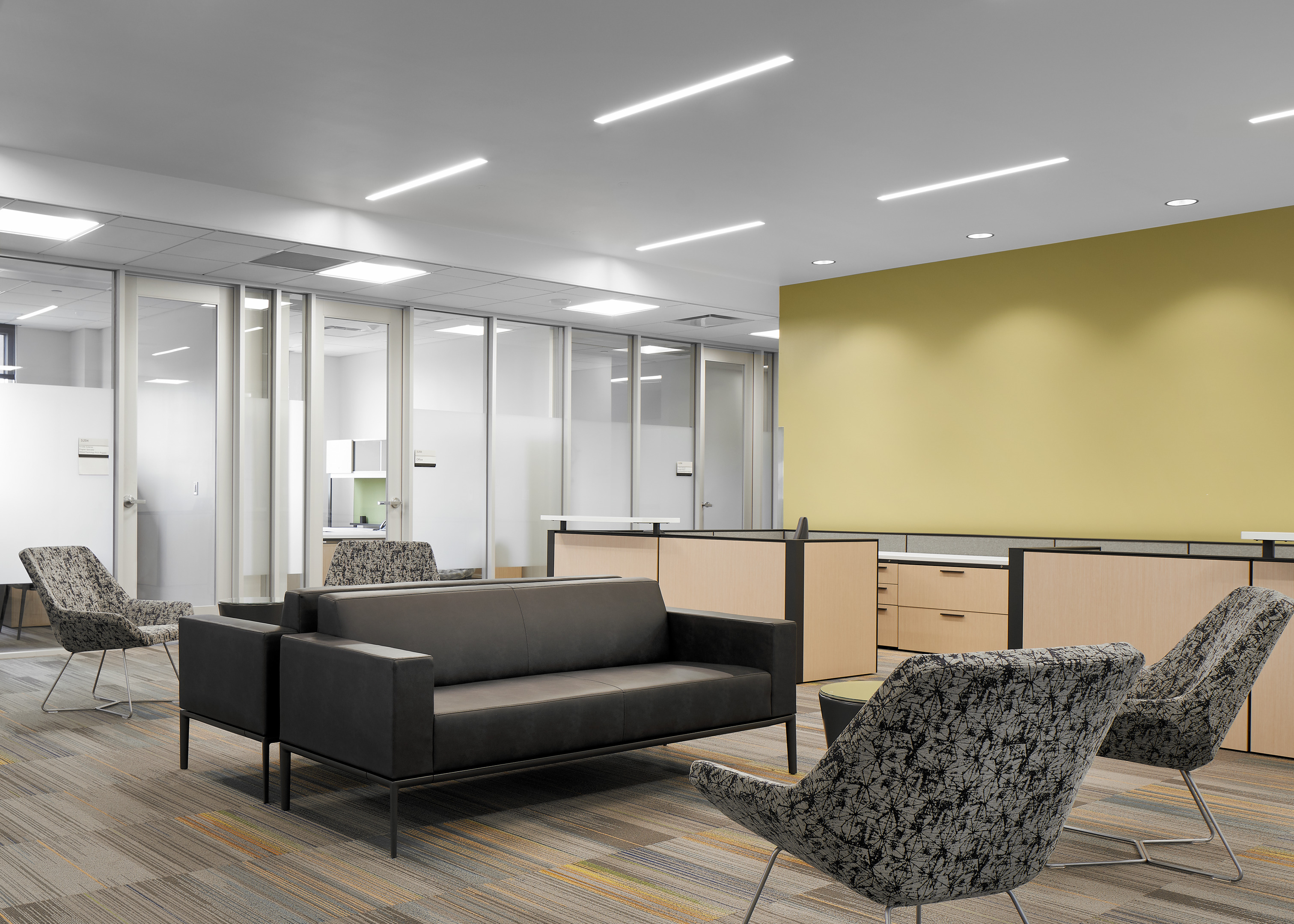 Modern office lobby with a black sofa, patterned armchairs, and neutral-toned walls. Fluorescent lighting above and partitioned office spaces in the background.