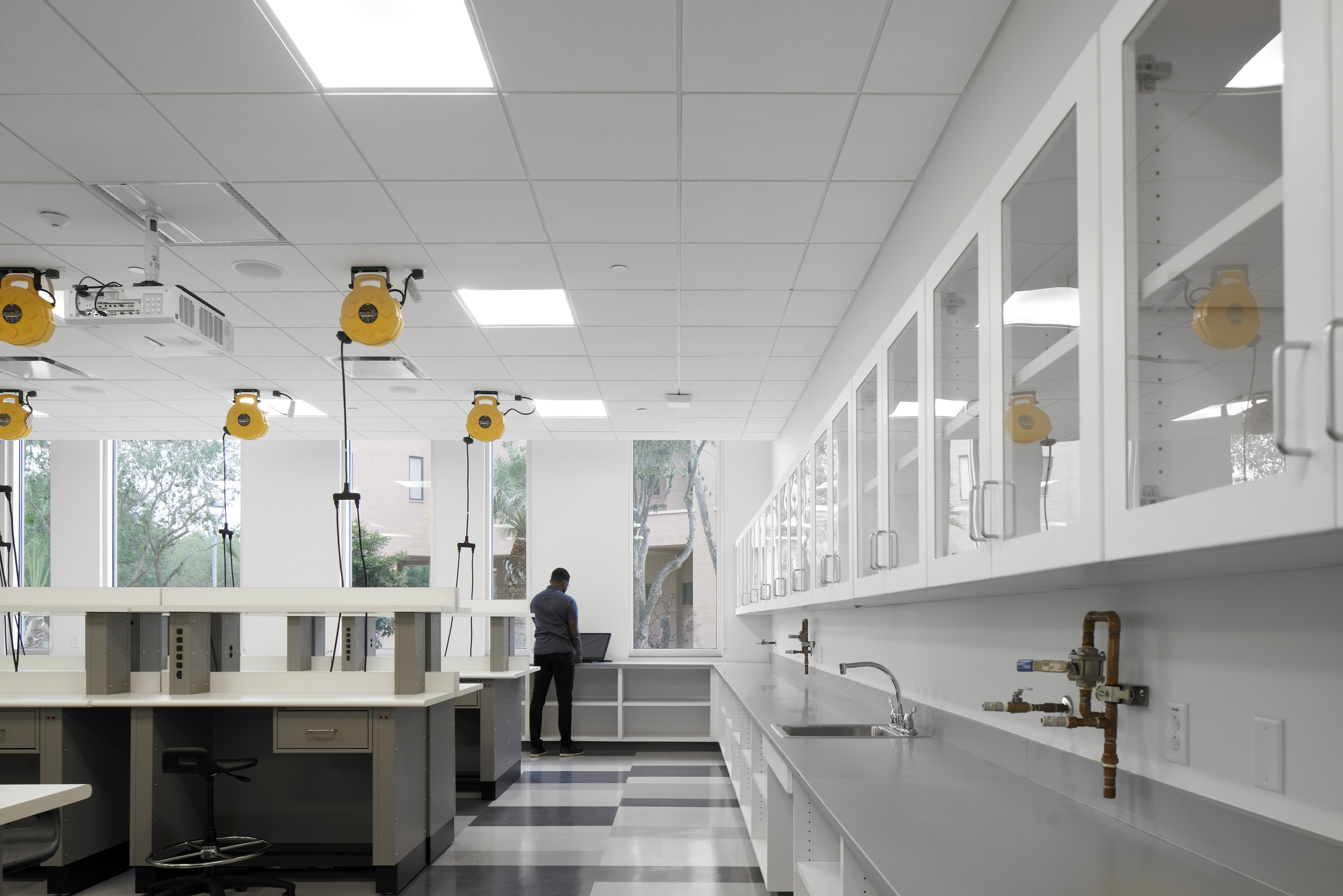 A person stands at a workstation in a modern, well-lit laboratory with white cabinets, countertops, and specialized equipment. Large windows allow natural light into the space.