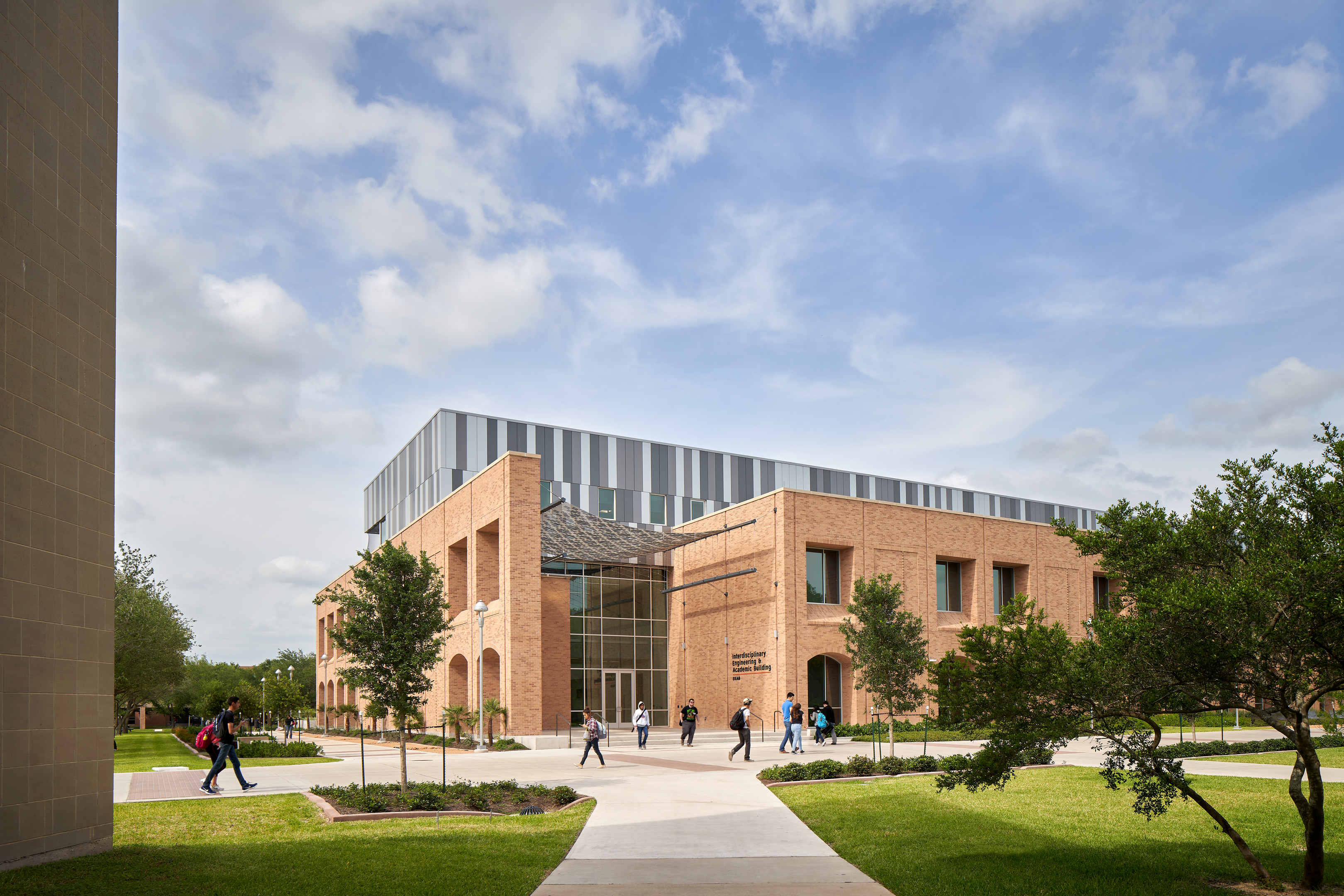 A modern brick academic building with large windows on a sunny day. Several people walk on the pathways leading to the building, surrounded by grass and trees.