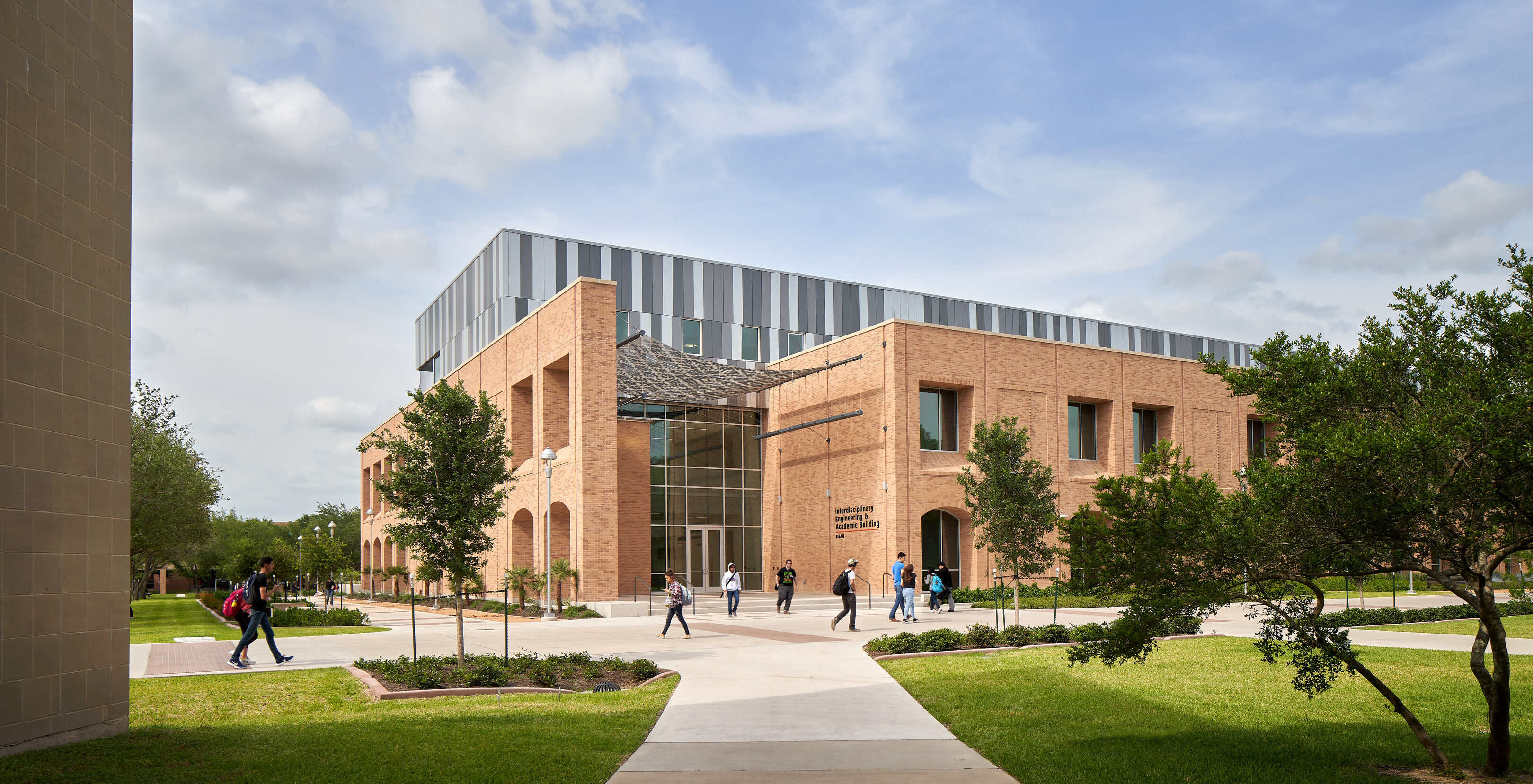 A modern brick university building with large glass windows under a partly cloudy sky stands proudly on the UTRGV Edinburg campus, surrounded by green lawns and trees. Several people walk along the pathways in front of this engineering hub.