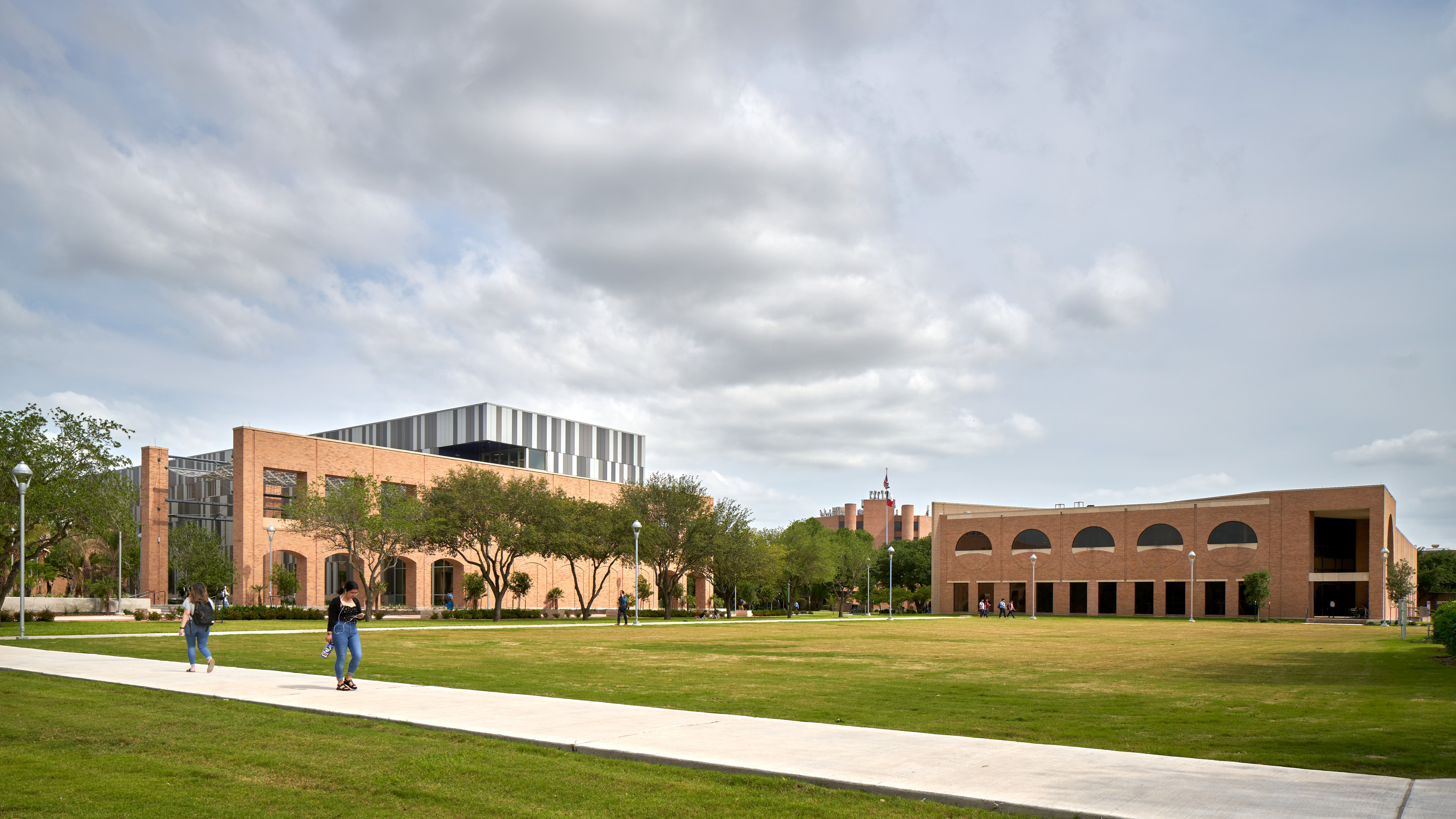 A spacious campus lawn at UTRGV Edinburg, bordered by modern brick buildings under a partly cloudy sky, with a few people walking along a white concrete path.