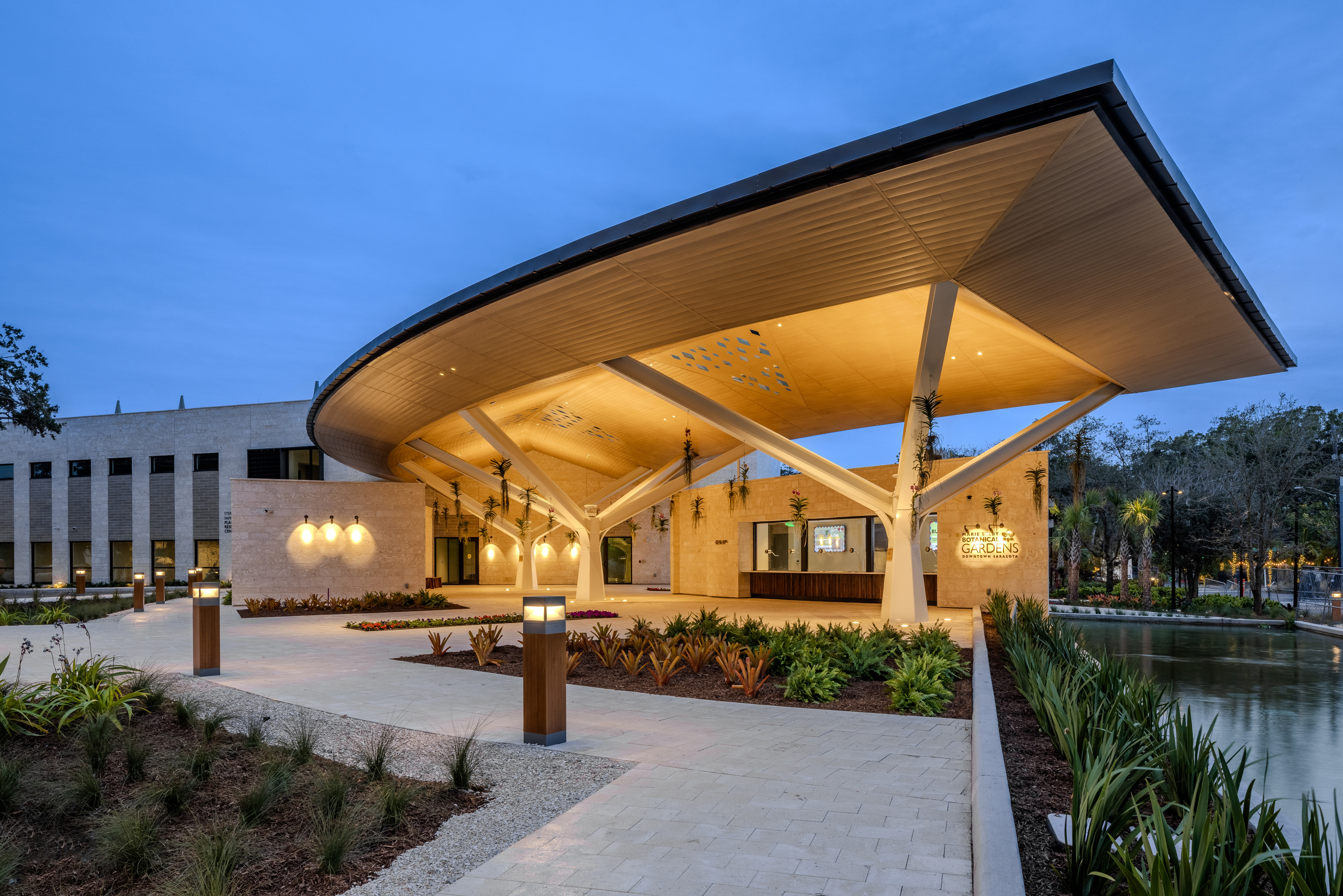 Modern building with a large, curved roof overhang supported by angled pillars, featuring landscaped gardens reminiscent of Selby Gardens and outdoor lighting. The building has multiple large windows and a sleek, contemporary design.