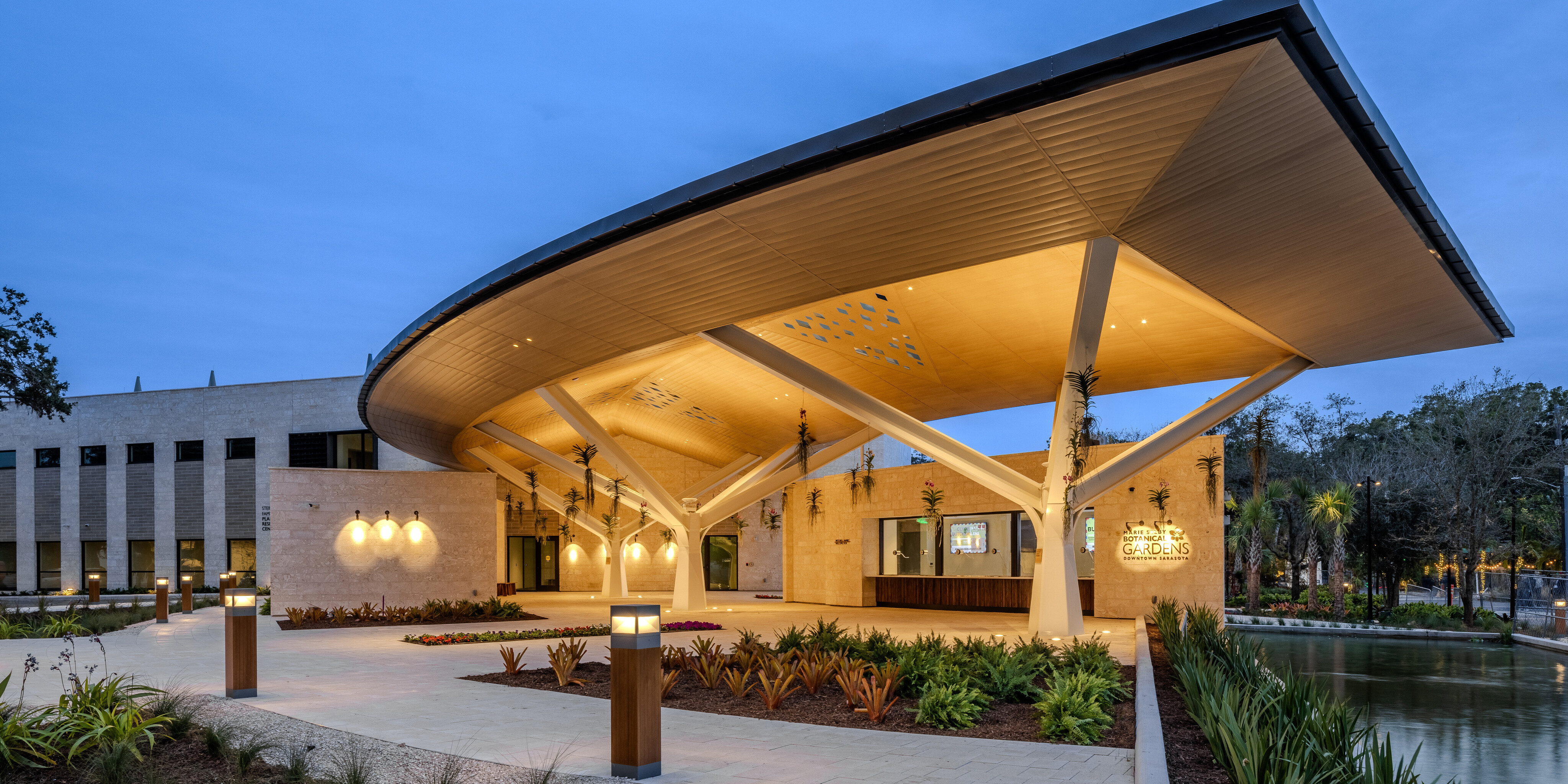 A modern building with a large, curved wooden roof and substantial supporting beams, featuring landscaped grounds with plants and pathway lights, stands gracefully adjacent to a waterway at dusk, reminiscent of the serene beauty found at Marie Selby Botanical Gardens.