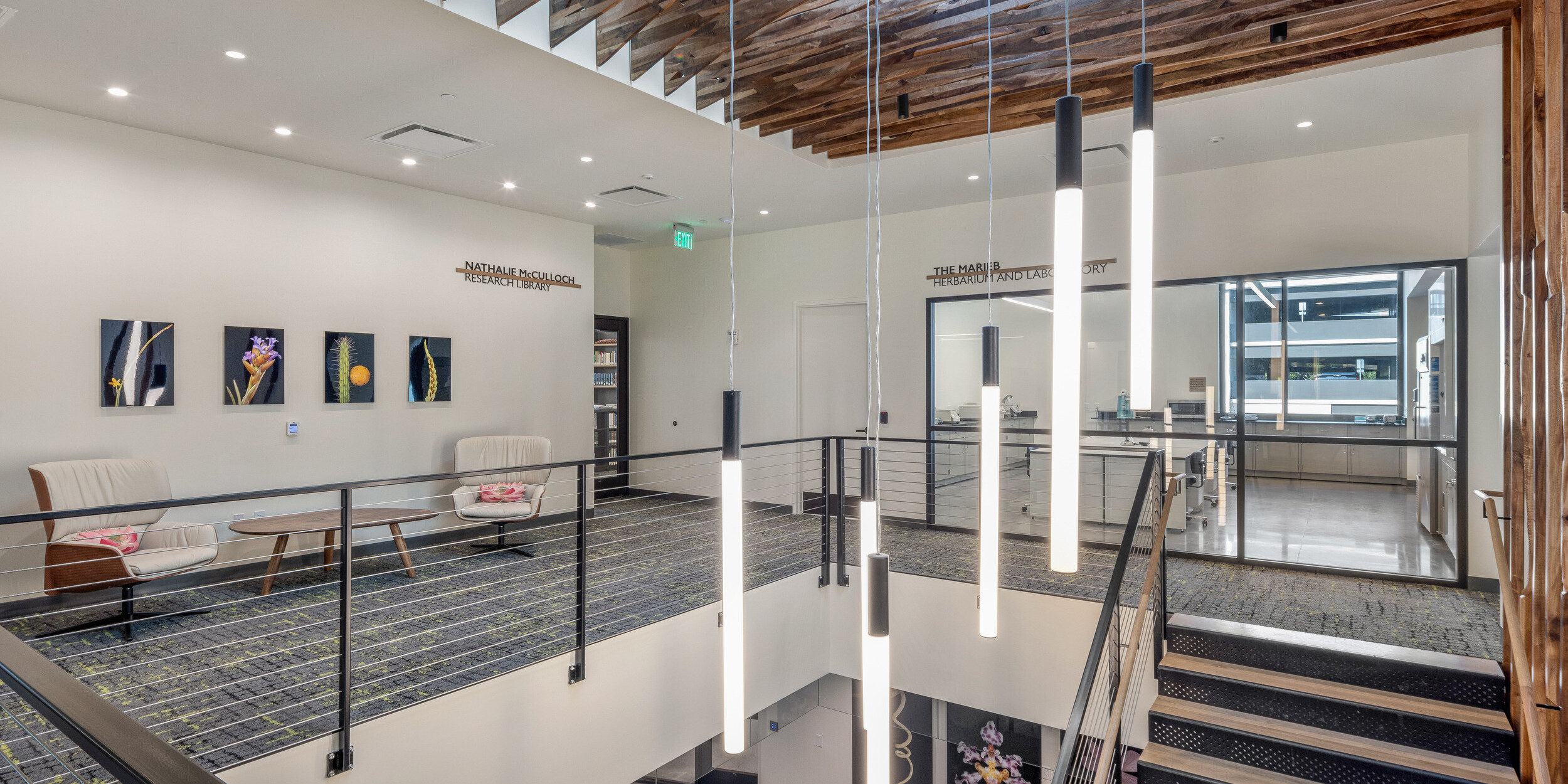 A modern interior space with a wooden ceiling, minimalist light fixtures, and a staircase with black railings. The walls feature framed artwork reminiscent of the elegance at Marie Selby Botanical Gardens, with office areas visible in the background.