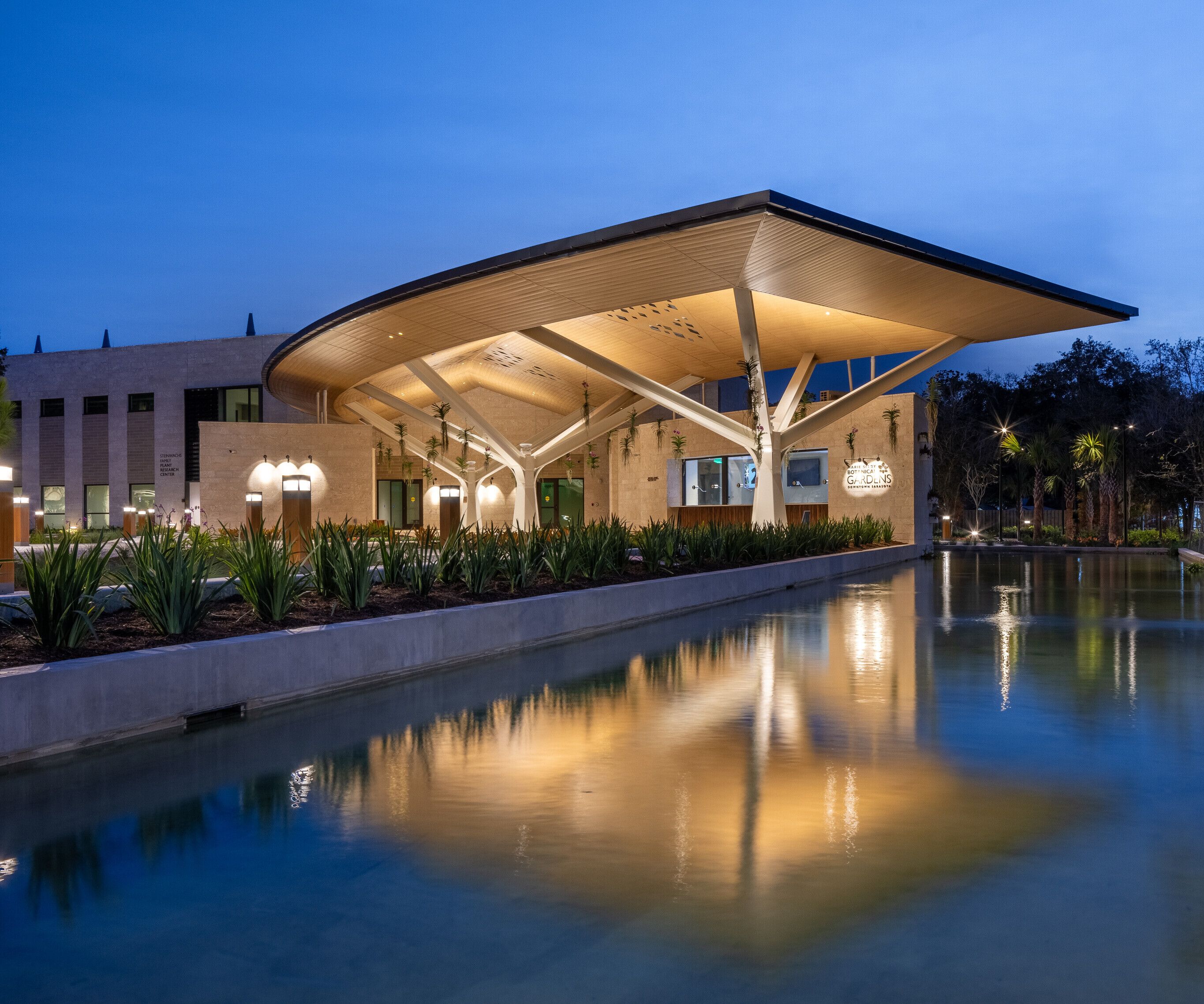 Modern building with a curved roof structure reflected in a rectangular water feature at dusk, reminiscent of the serene beauty found at Marie Selby Botanical Gardens.