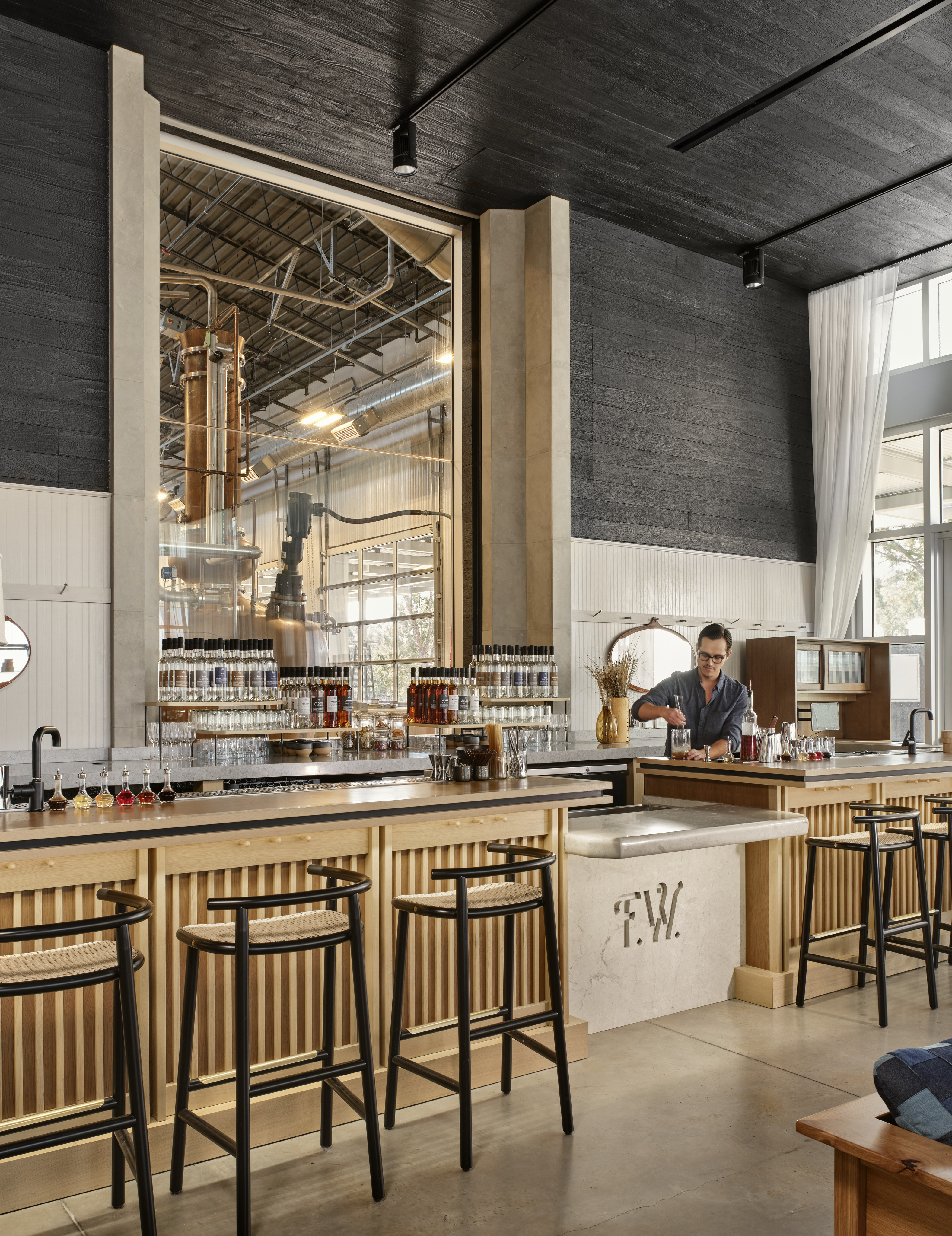 A man works behind a modern, well-lit bar with a variety of bottles displayed in front and large distillation equipment visible through a large window in the background. The atmosphere exudes the meticulous craftsmanship of Fierce Whiskers Distillery.