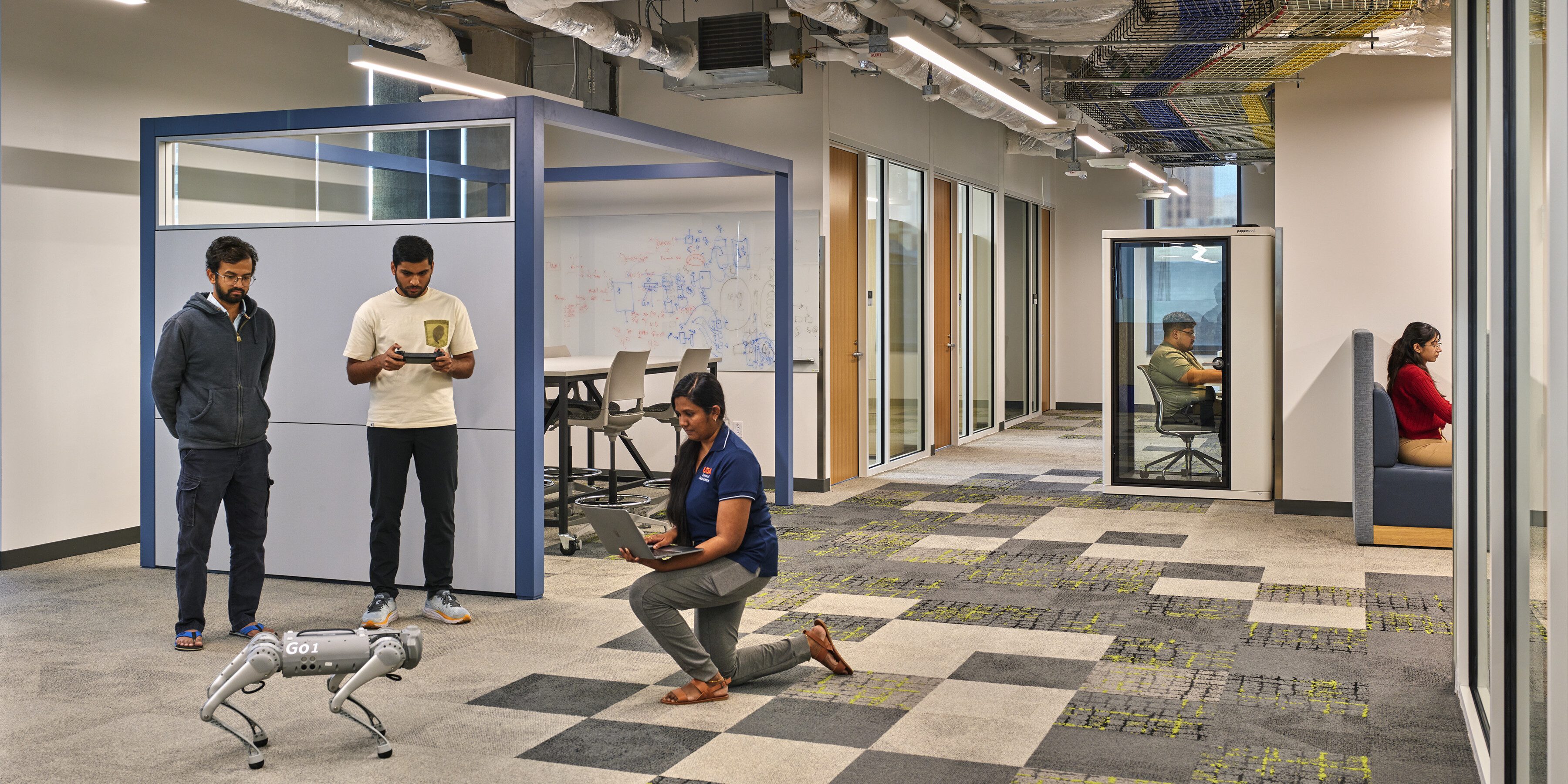 Three men observe and interact with a robot dog in an office space with exposed ceilings and modern furnishings at San Pedro I in San Antonio; a person is seated in a glass-walled booth while another is working in a private alcove.