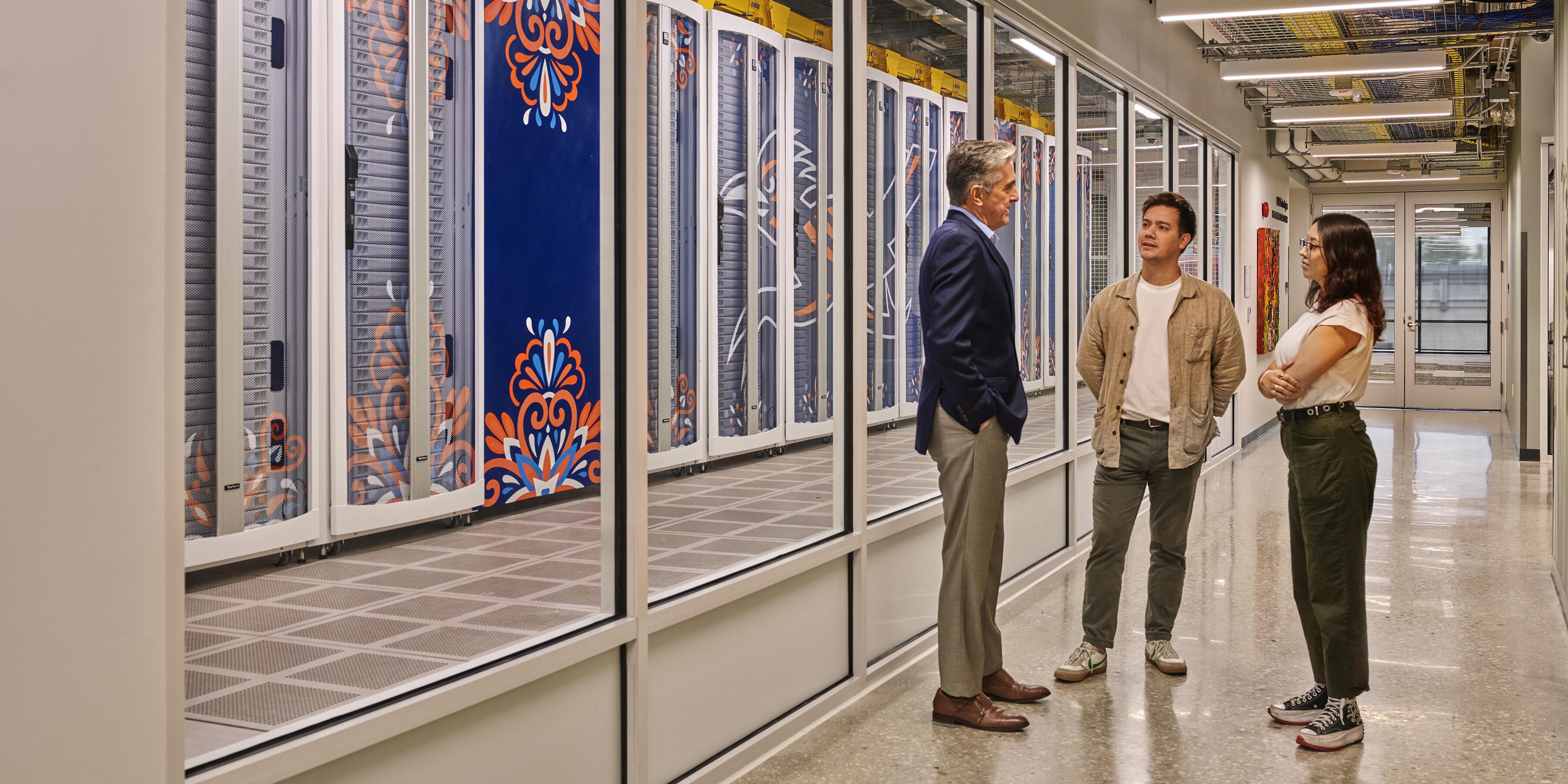 Three people stand conversing in a corridor next to a glass wall with a view of server racks inside San Pedro I, the cutting-edge data center at the University of Texas at San Antonio (UTSA).