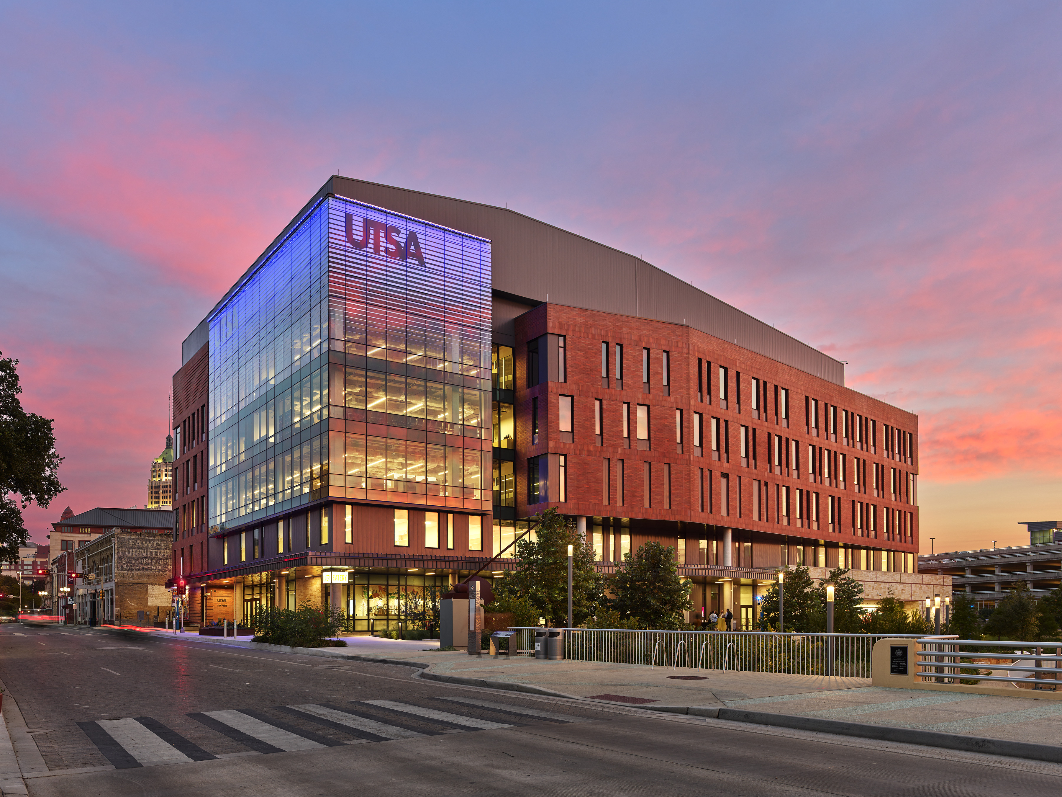 Modern five-story building with large glass windows and "UTSA" signage lit up near the top. The sky is a vibrant mix of pink and orange during sunset, perfectly capturing San Antonio's charm. San Pedro I stands proudly, with a road and lush greenery in the foreground.