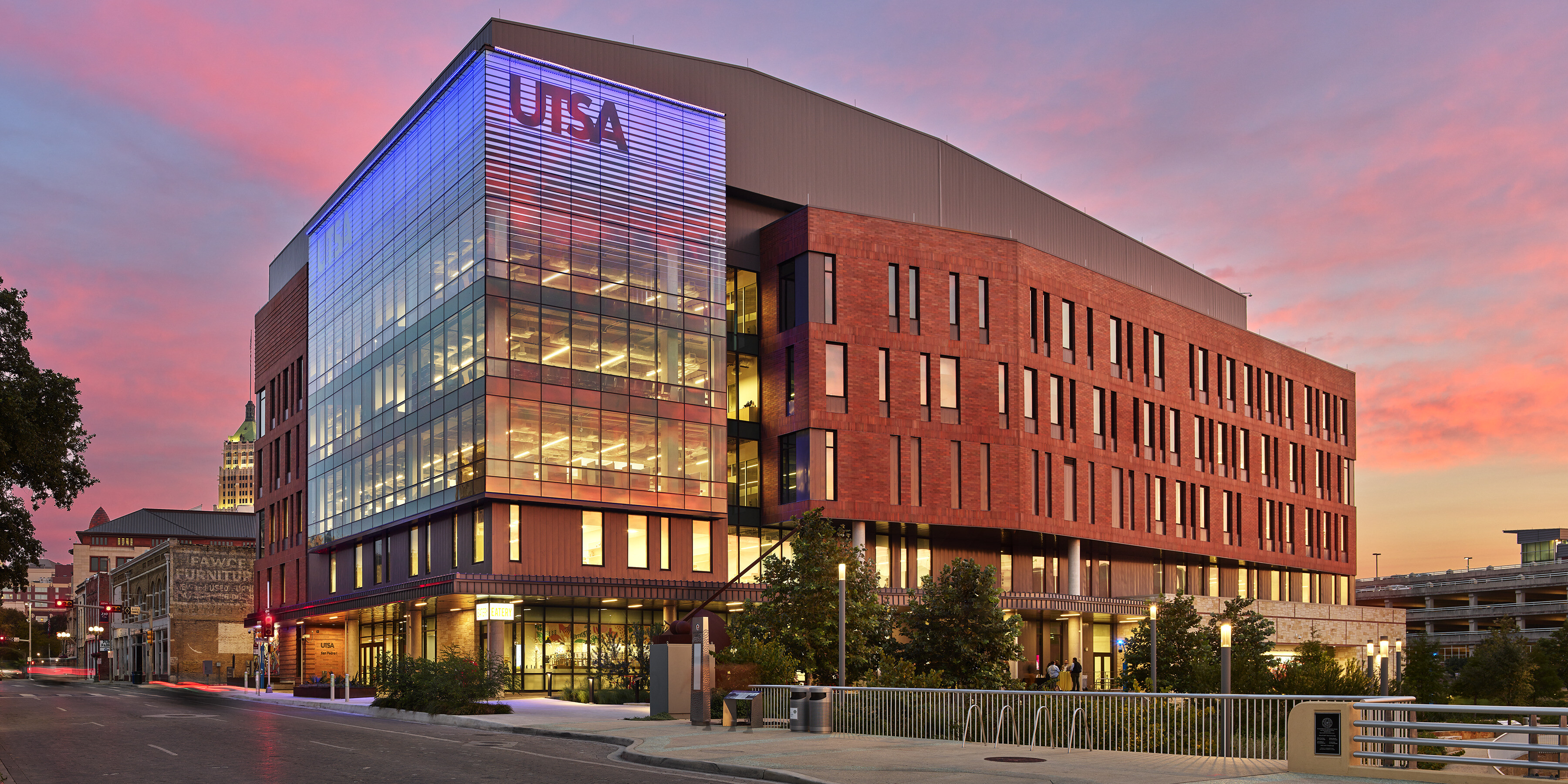 A modern building with "UTSA" on its facade, featuring glass and brick elements, is illuminated at dusk under a pink and blue sky in San Antonio. This impressive structure represents the University of Texas's San Pedro I campus.