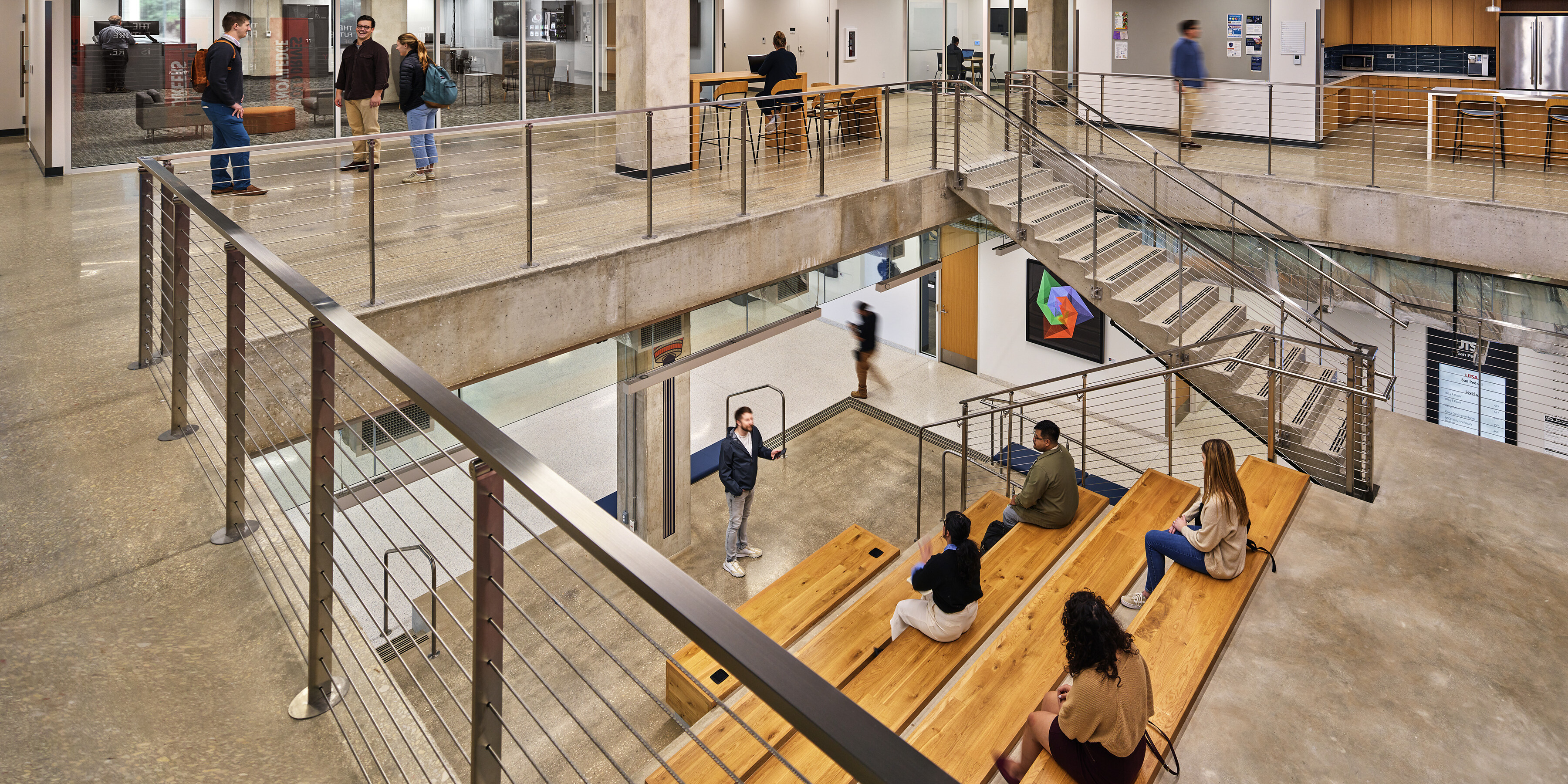 A modern, open-concept multi-level office space with people working and socializing. The San Pedro I area features concrete floors, glass walls, a staircase, and wooden seating areas, reminiscent of the innovative spaces at the University of Texas at San Antonio.