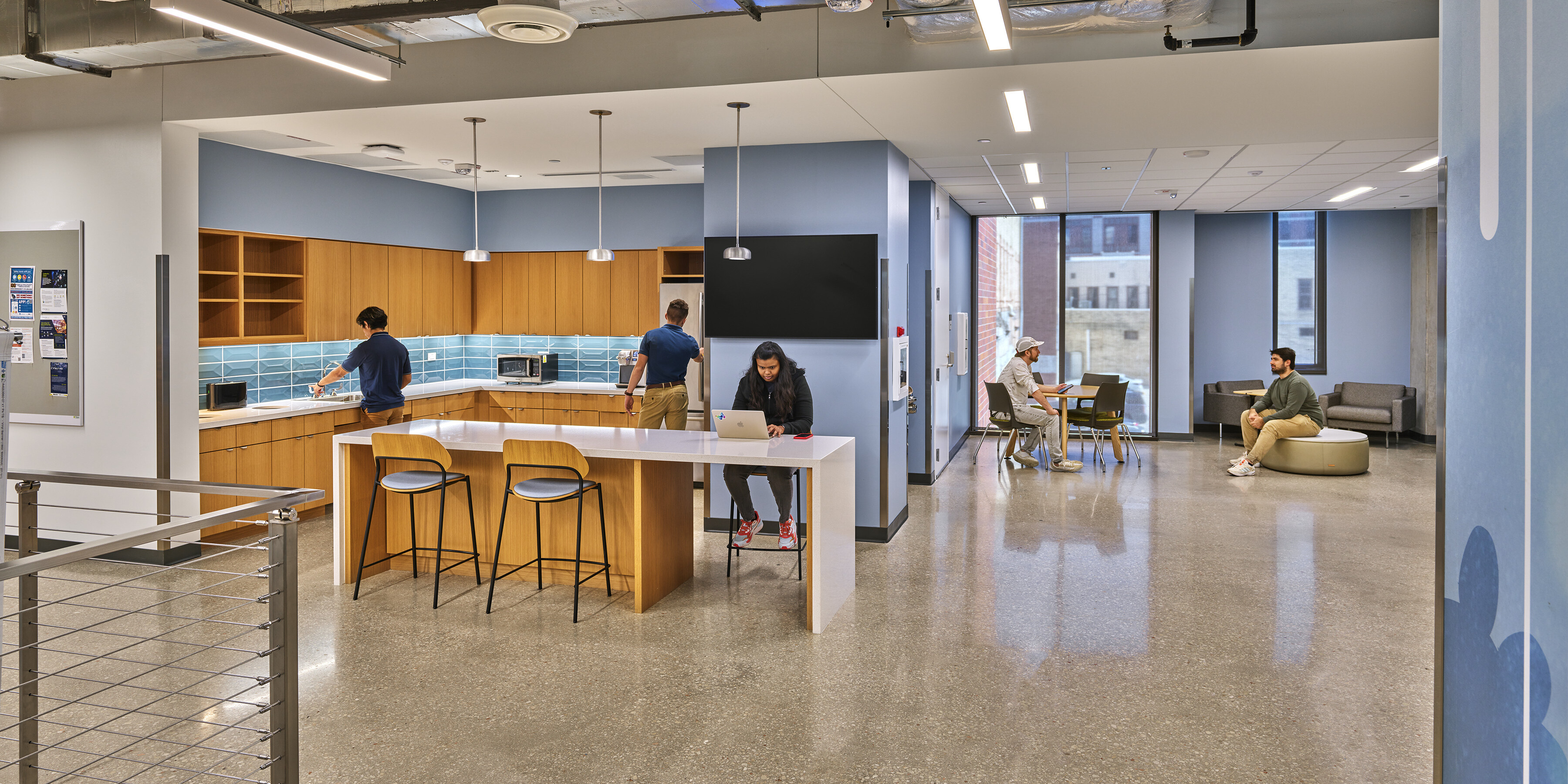 Modern office break room with people working at tables and counters; amenities include a kitchenette with blue backsplash, microwave ovens, seated individuals using laptops, and an exposed ceiling. Located in the San Pedro I building near the University of Texas at San Antonio.