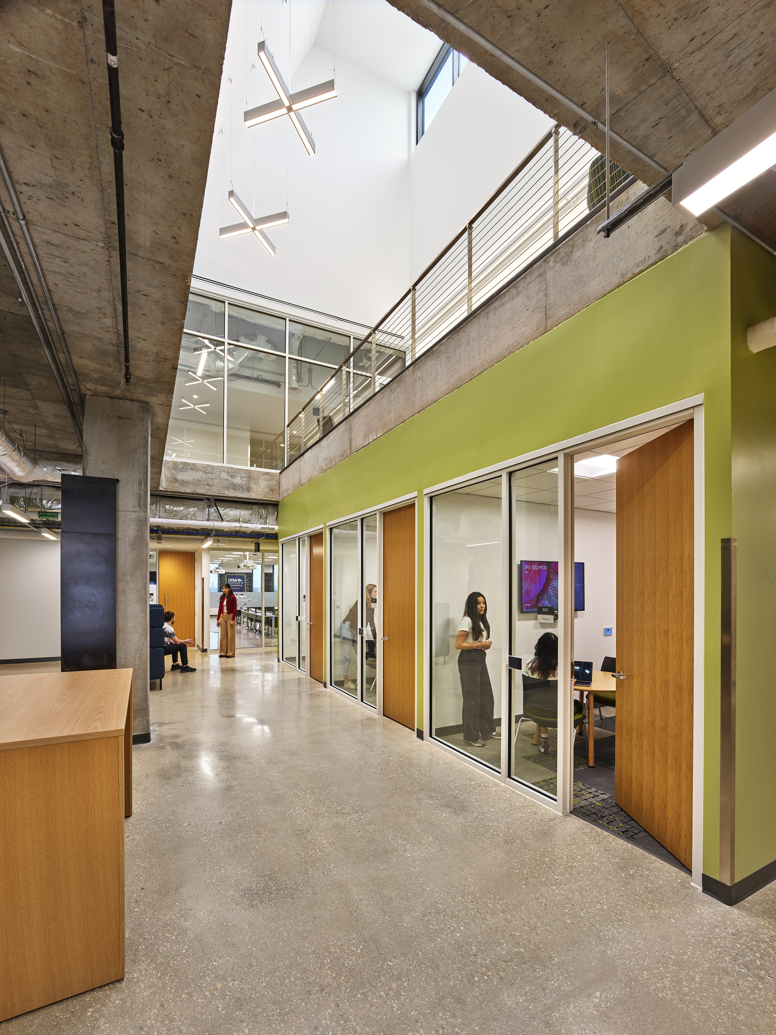 A spacious, modern office interior at San Pedro I with glass-walled meeting rooms on the right and an open seating area on the left. Individuals are seen working and conversing inside the rooms and in the hallway, fostering a vibrant atmosphere similar to that of University of Texas, San Antonio.