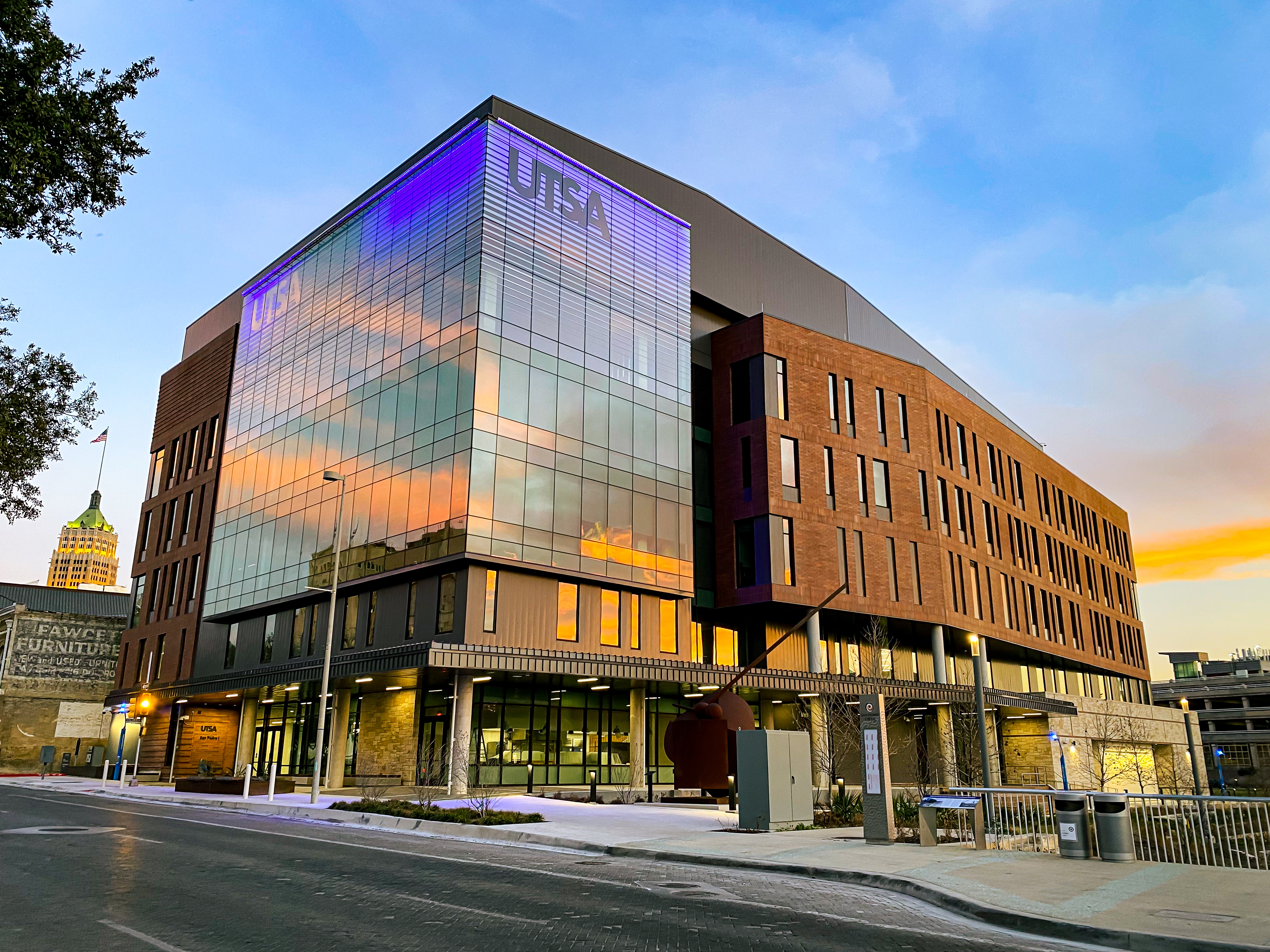 A modern, multi-story building with a glass and brick exterior showcases the glowing UTSA logo near the top, reflecting the San Antonio sunset sky. Known as San Pedro I, this striking structure is bordered by a sidewalk and street.