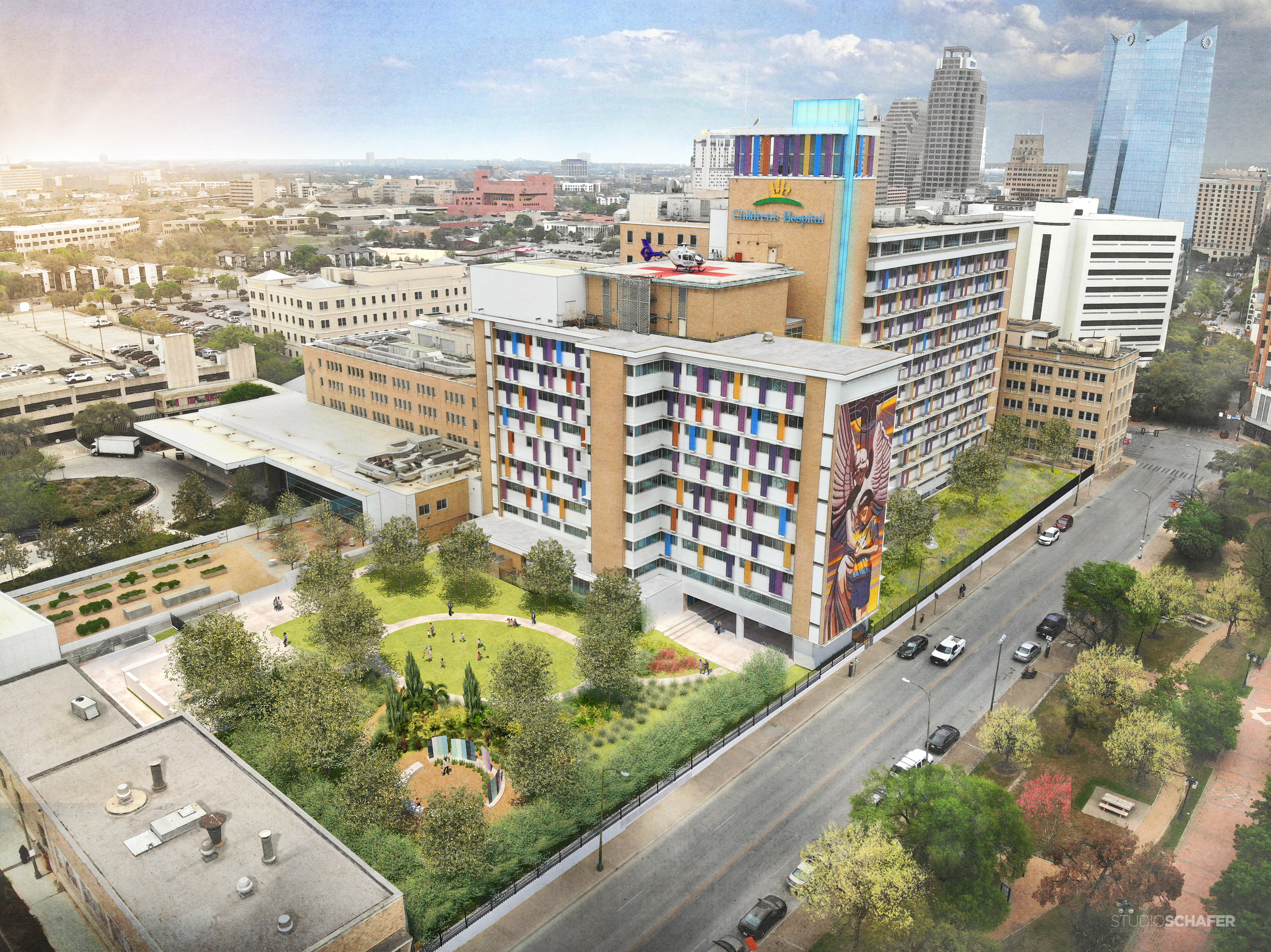 Aerial view of Christus Children's Hospital in San Antonio, a multistory, colorful building adorned with murals and surrounded by Biophilic Gardens, trees, and adjacent structures in a city landscape.