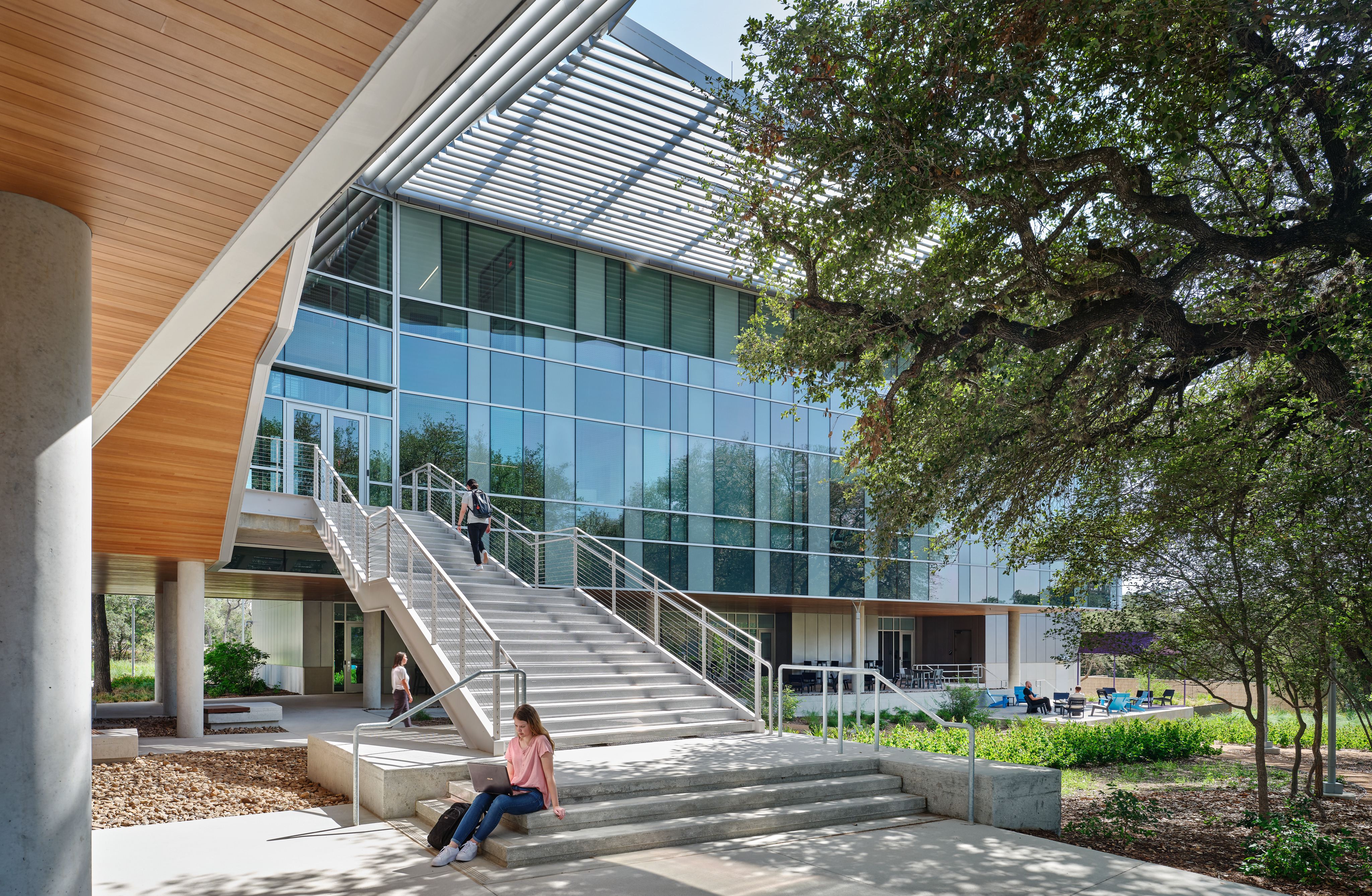 A modern building with large windows and an exterior staircase at Northwest Vista College. A person sits on the steps reading, and others are visible in the background. The area is shaded by trees and the building's overhang, part of the campus' renowned STEM Center under Alamo Colleges.