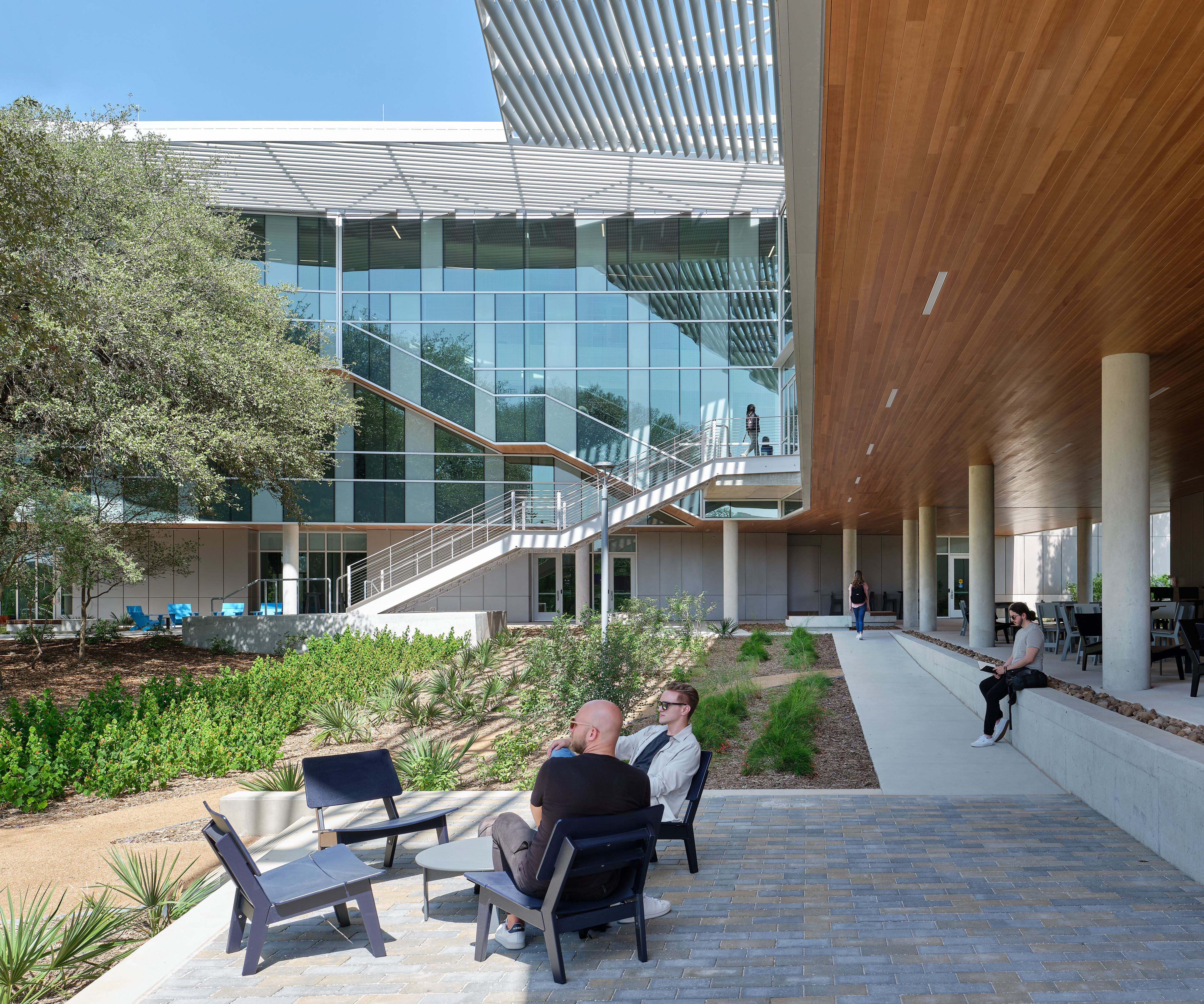 People sit on chairs in a courtyard with modern architectural elements including a glass facade, wooden ceiling, and landscaped greenery at the STEM Center of Northwest Vista College. Stairs lead to an upper level of the building.