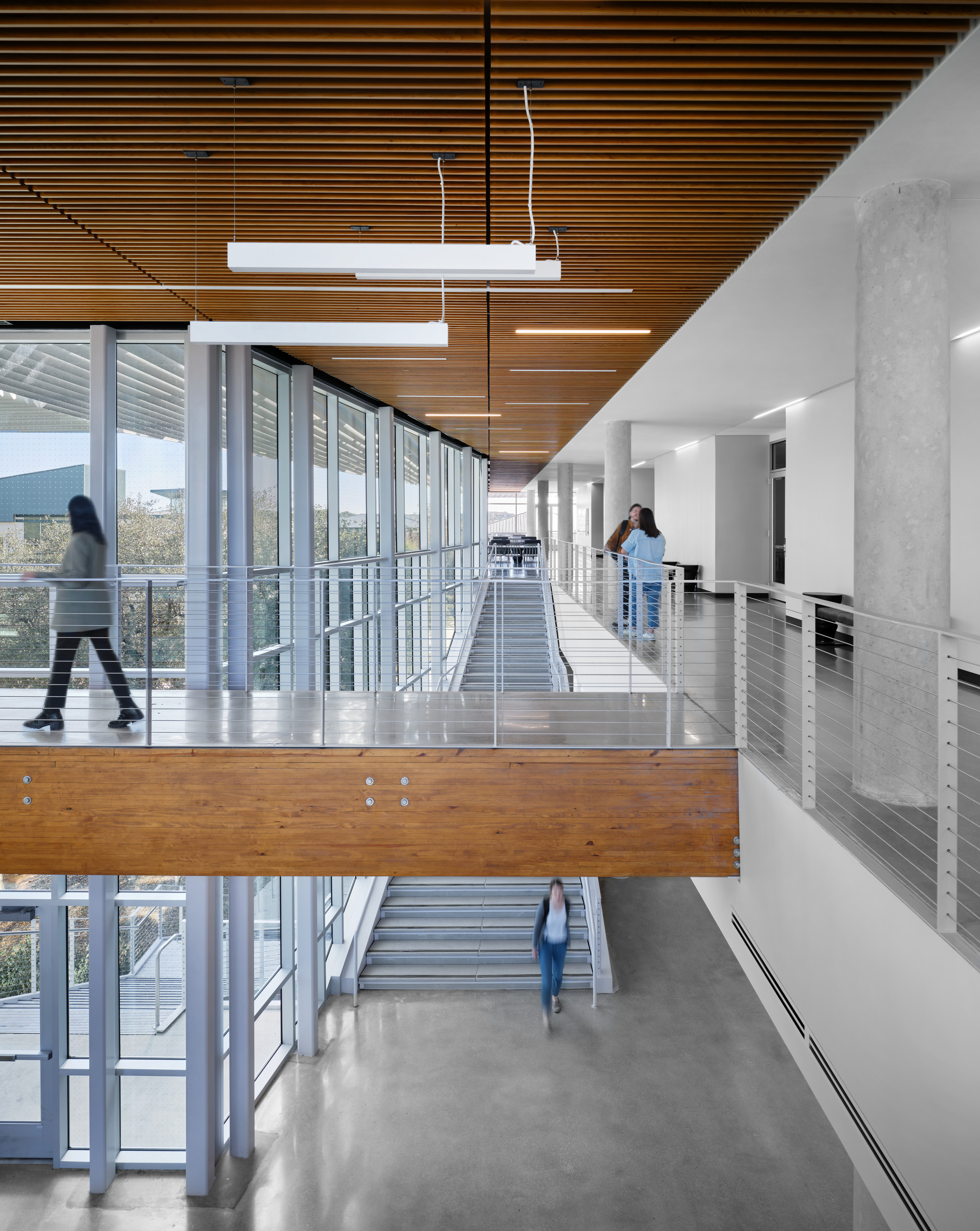 Inside a modern building at Northwest Vista College, multiple people walk across a spacious, well-lit upper walkway with wooden ceiling and glass railings, overlooking a staircase and an open area below—a vibrant glimpse into the bustling STEM Center at Alamo Colleges.