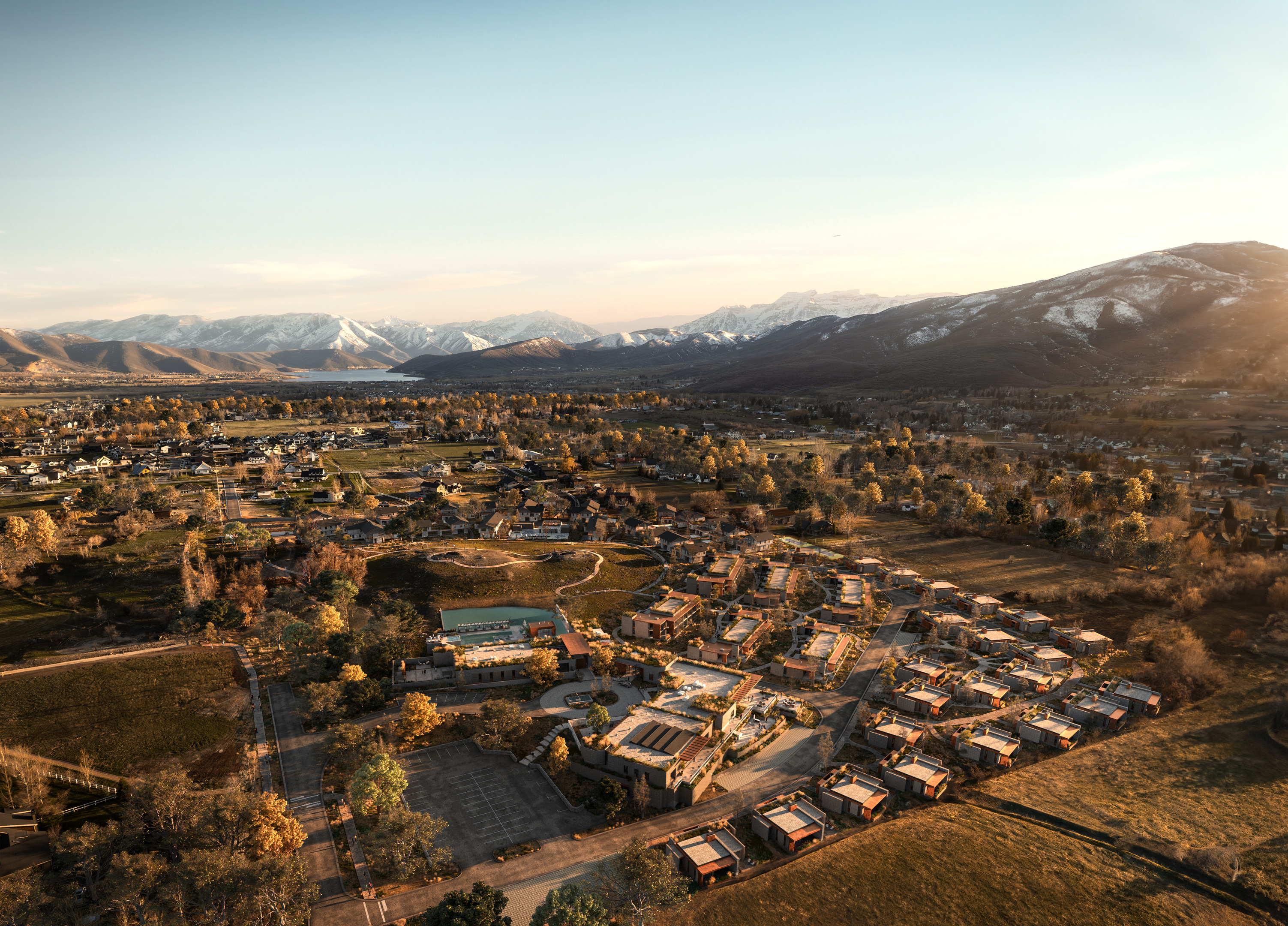 Aerial view of a residential area with houses and buildings surrounded by fields and trees, with snow-capped mountains and a lake visible in the background under a clear sky, near the serene AMEYALLI Resort.