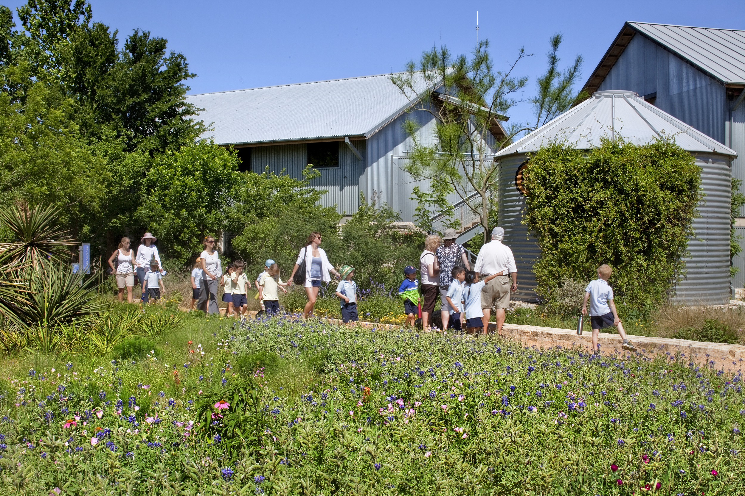 A group of people walks alongside a garden filled with various flowers at the Lady Bird Johnson Wildflower Center, near a metal tank and buildings, on a sunny day.