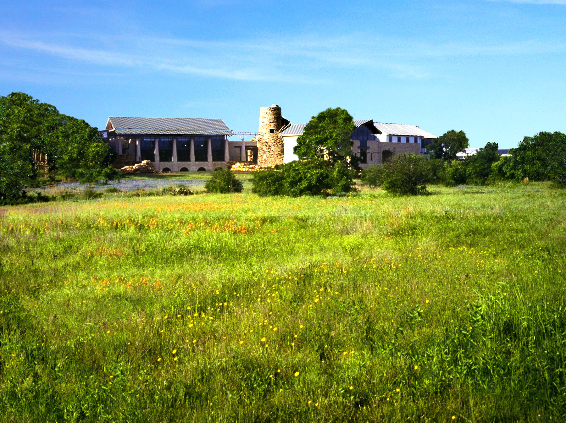 A large stone building with a metal roof stands majestically in the middle of the green grassy field at the Wildflower Center, surrounded by scattered trees on a clear, sunny day.