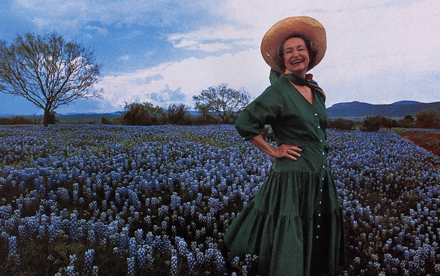 Woman in a green dress and straw hat standing in a field of bluebonnets, smiling with hands on hips. Trees and mountains are visible in the background under a partly cloudy sky, reminiscent of the fields at the Lady Bird Johnson Wildflower Center.