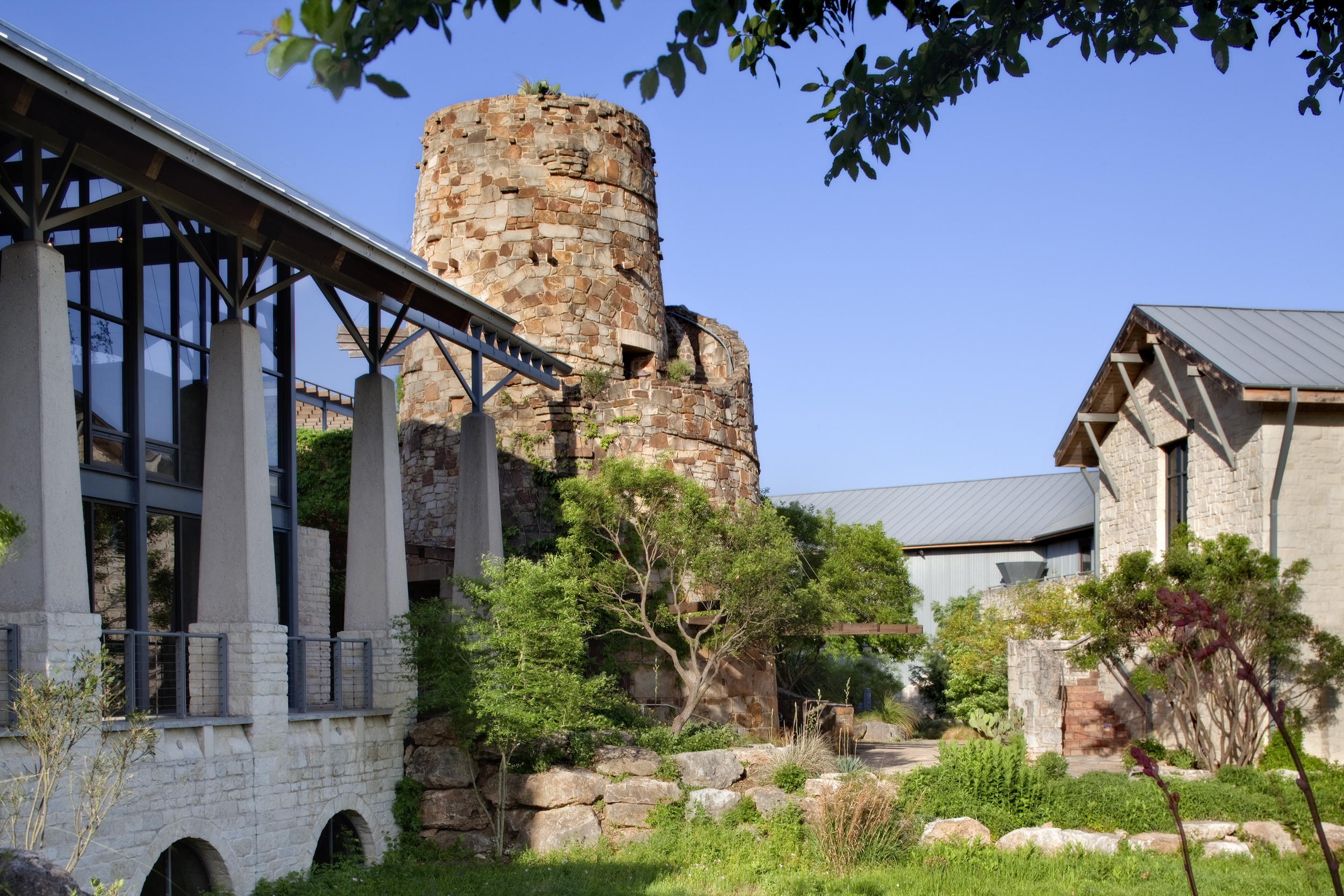 A stone tower and modern buildings surrounded by greenery under a clear blue sky evoke the charm of the Lady Bird Johnson Wildflower Center.