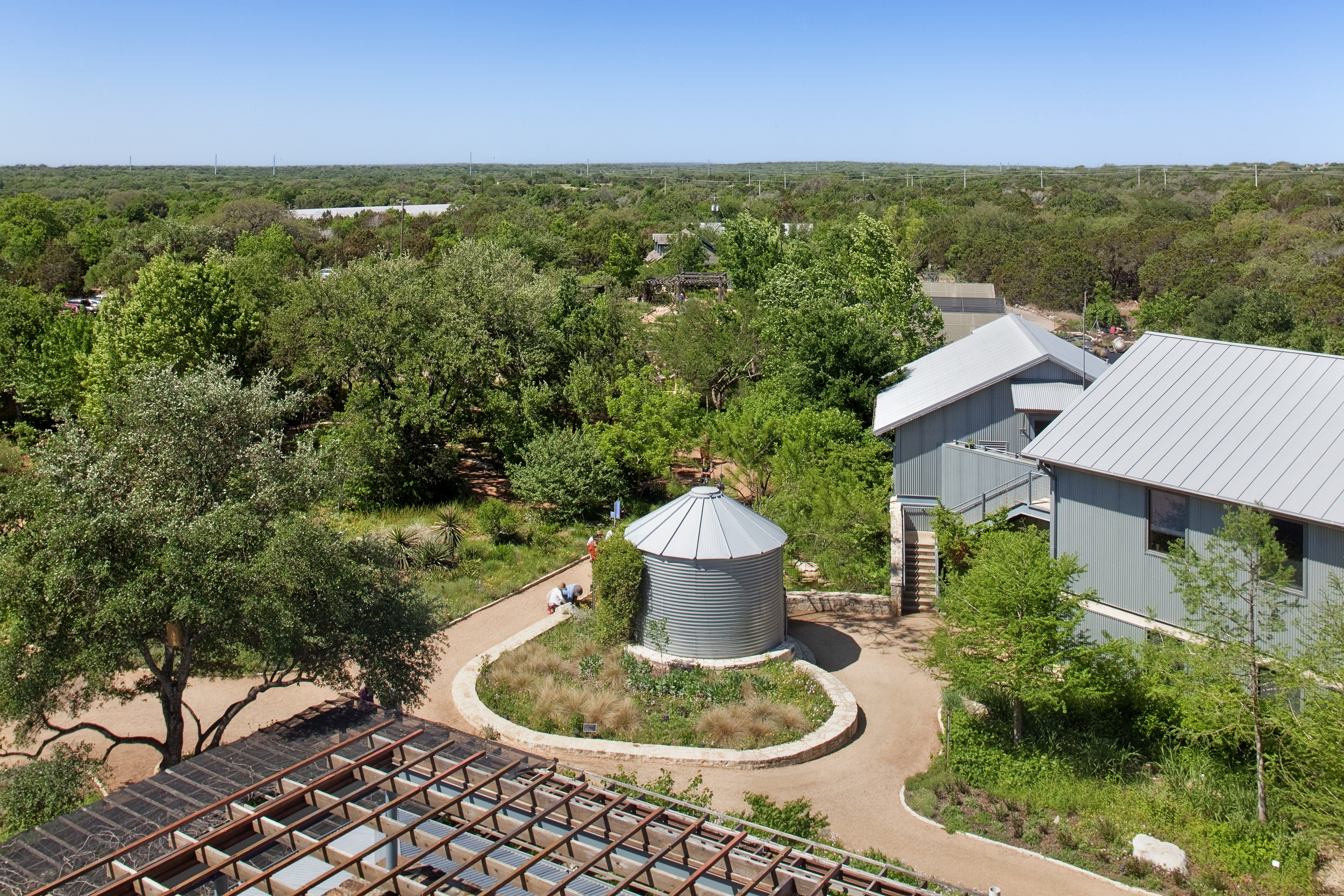 Aerial view of the Lady Bird Johnson Wildflower Center's green-roofed building and a cylindrical structure, surrounded by lush greenery and a winding path on a bright, clear day.