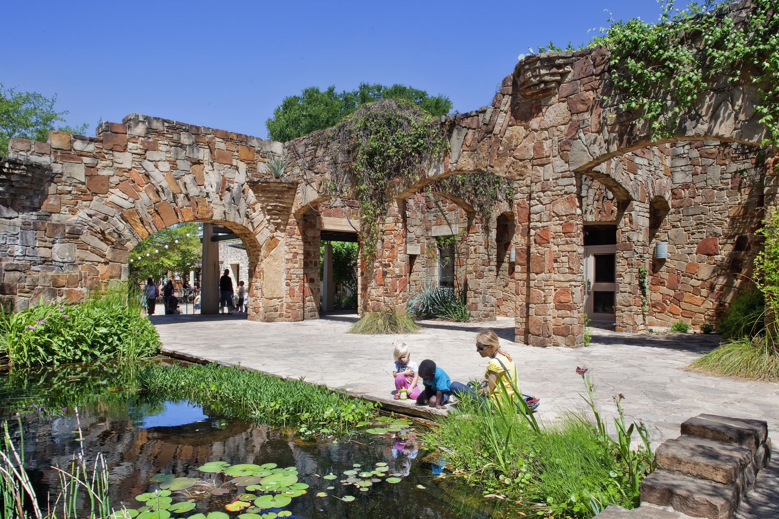 A stone structure with archways and climbing plants stands near a small pond with lily pads at the Lady Bird Johnson Wildflower Center. A woman and two children are seated at the pond's edge under a bright blue sky.