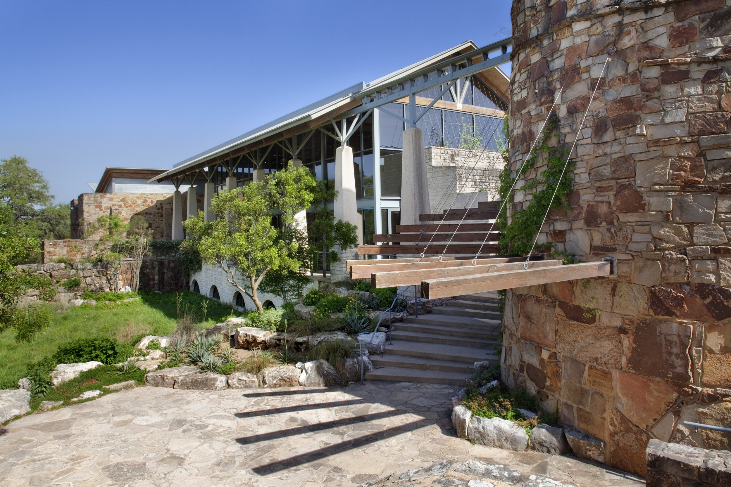 A modern building with large glass windows and a stone facade, featuring a suspended wooden bridge and surrounded by a landscaped garden reminiscent of the Lady Bird Johnson Wildflower Center.