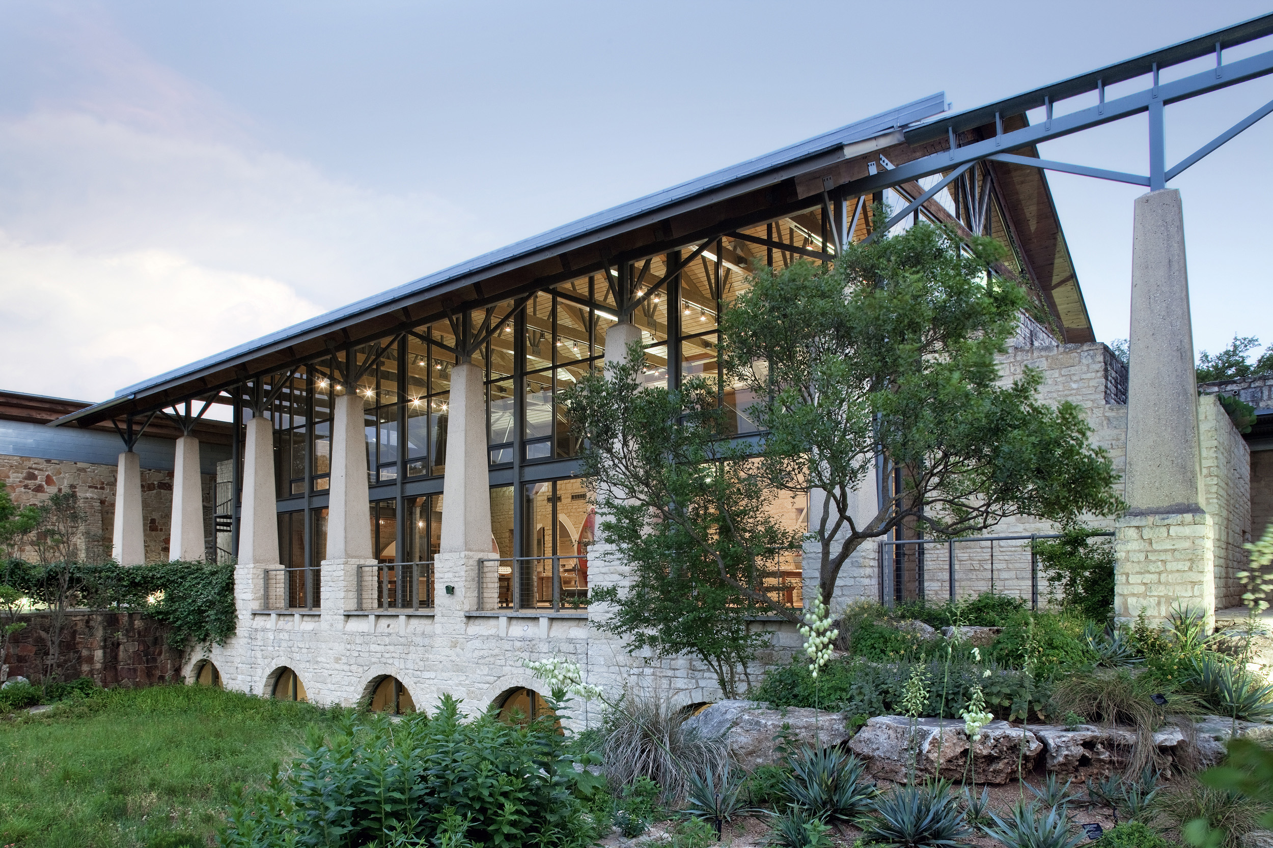 A modern building with large glass windows and stone pillars, reminiscent of the architecture at the Lady Bird Johnson Wildflower Center, surrounded by lush greenery and vibrant plants.