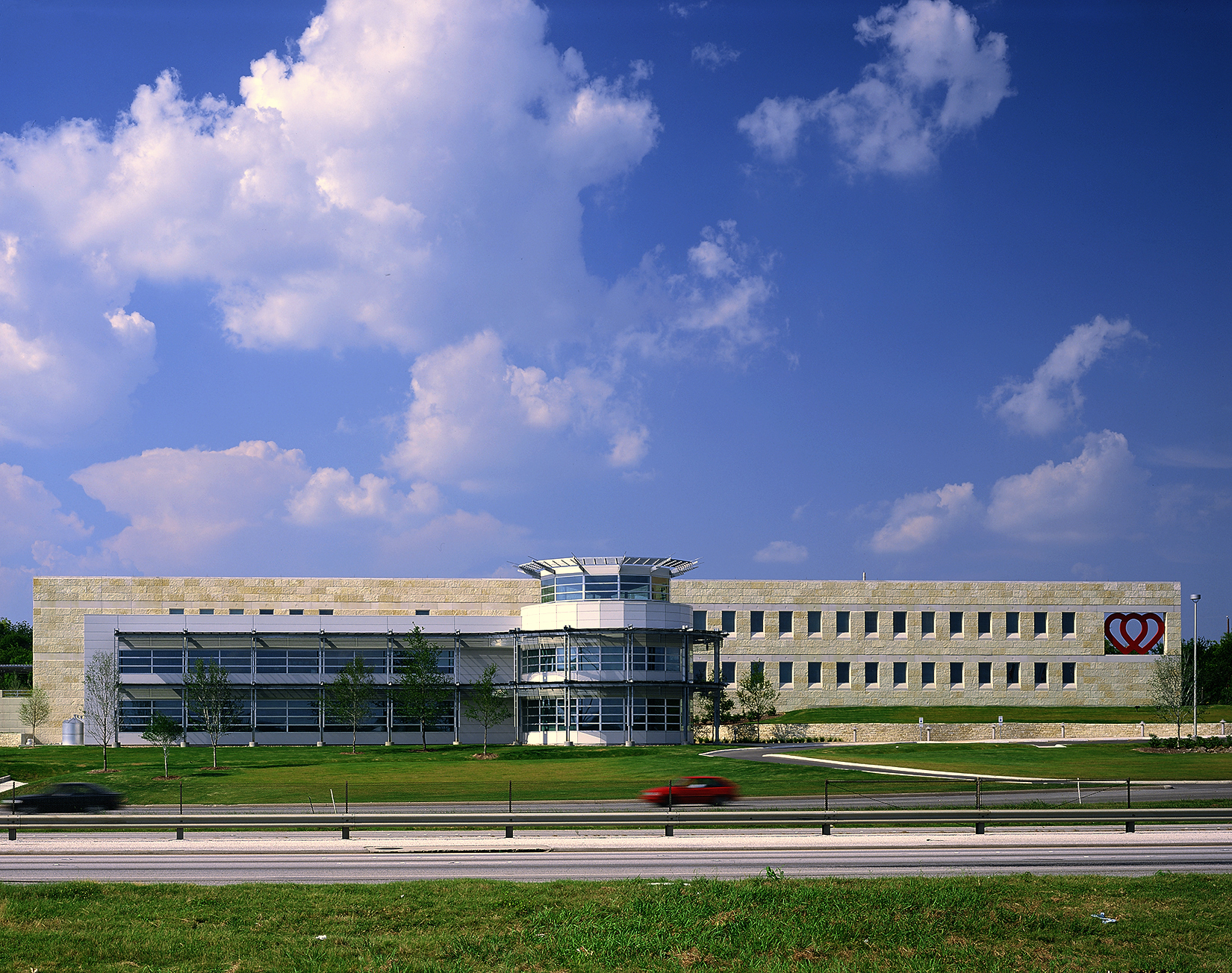 A modern, rectangular office building with a heart logo on the right side, set against a partly cloudy sky, houses the South Texas Blood and Tissue Center. Two cars drive along the road in front of the building.