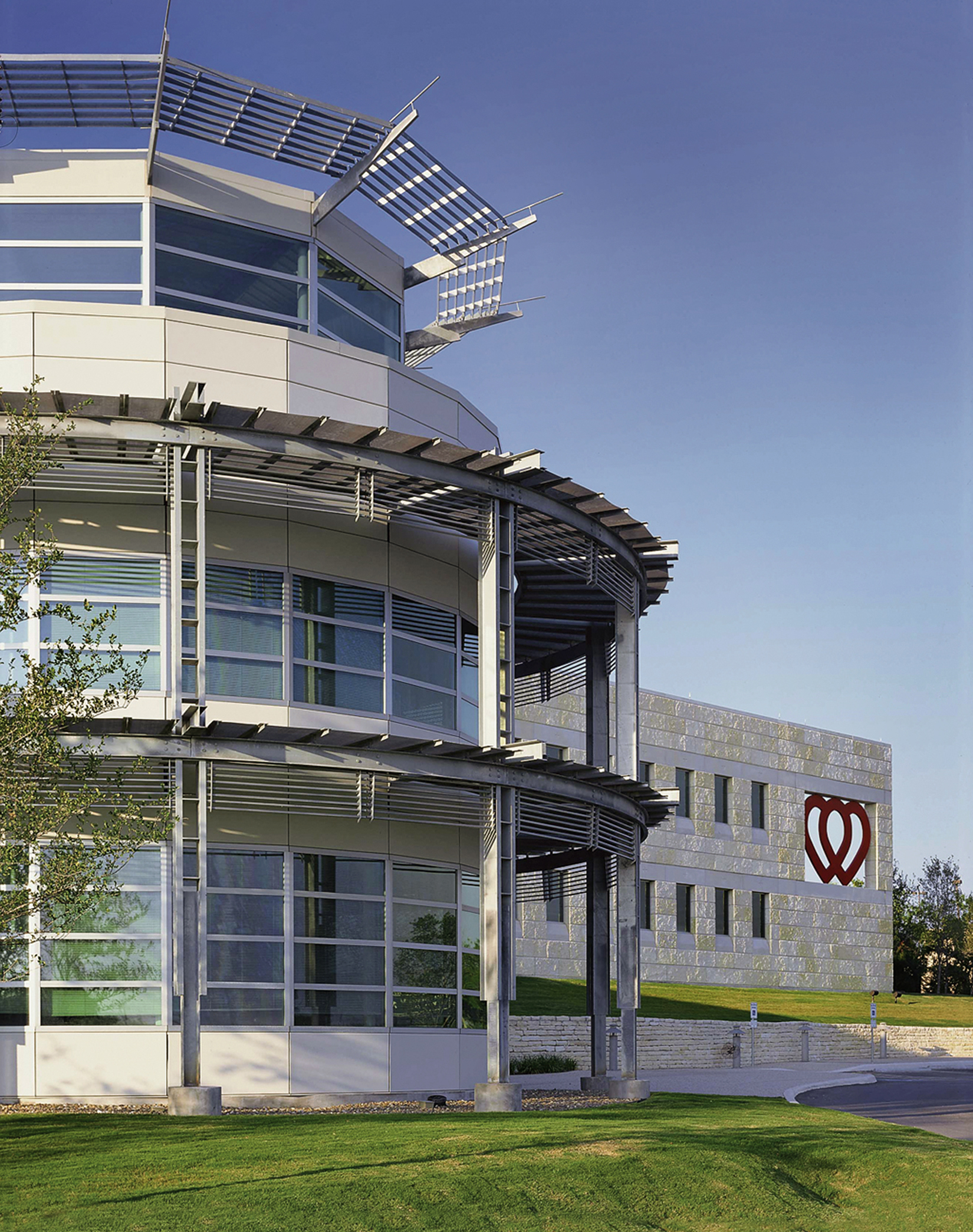 Modern multi-story building with large windows and metallic accents under a clear sky. A building with a heart-shaped logo of the South Texas Blood and Tissue Center is visible in the background. Green lawn in the foreground.