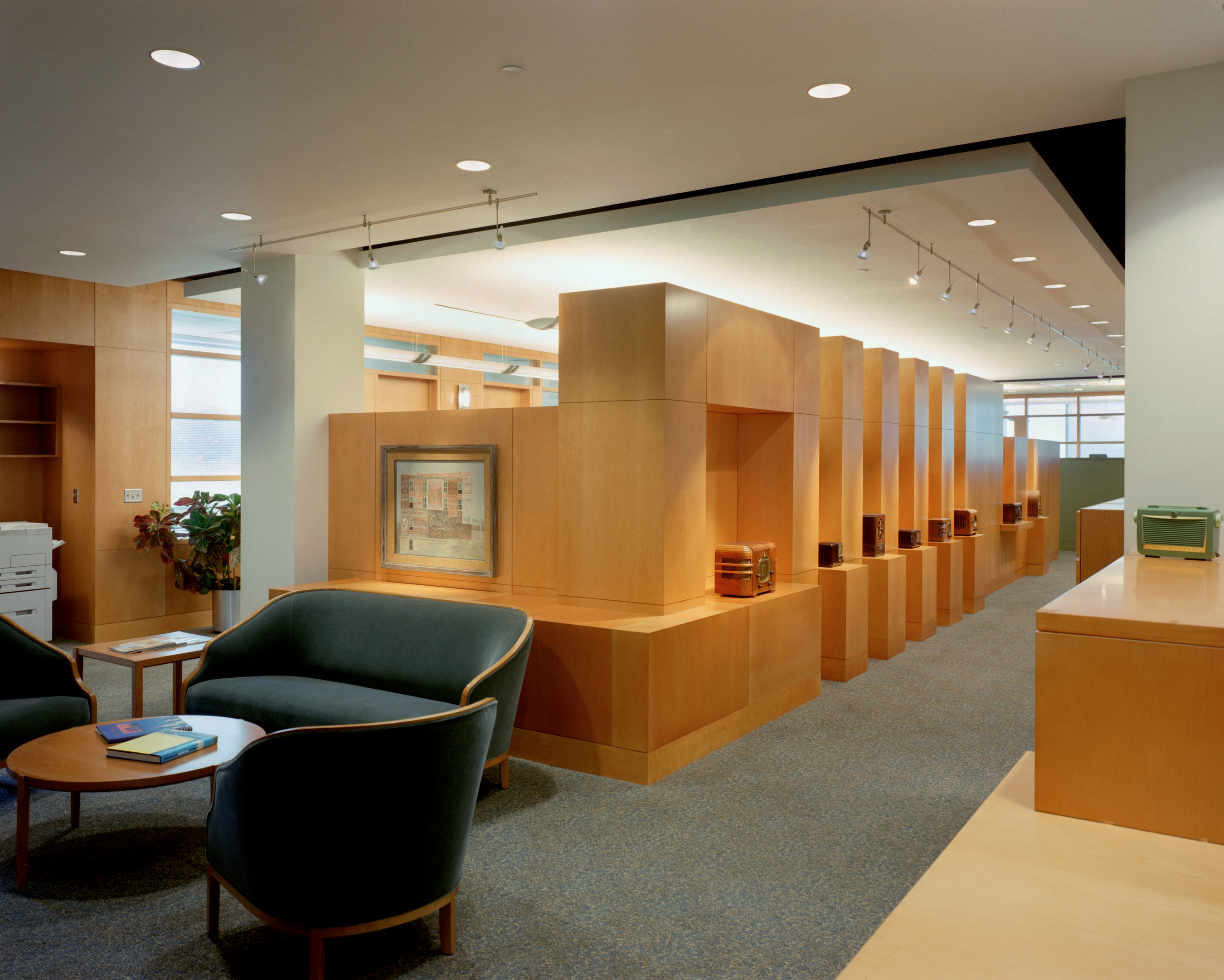 The headquarters' modern office interior boasts wooden furniture, a cozy seating area with chairs and a table, and a display wall showcasing books and framed art. Clear Channel's influence is evident throughout, with overhead lighting beautifully illuminating the space.