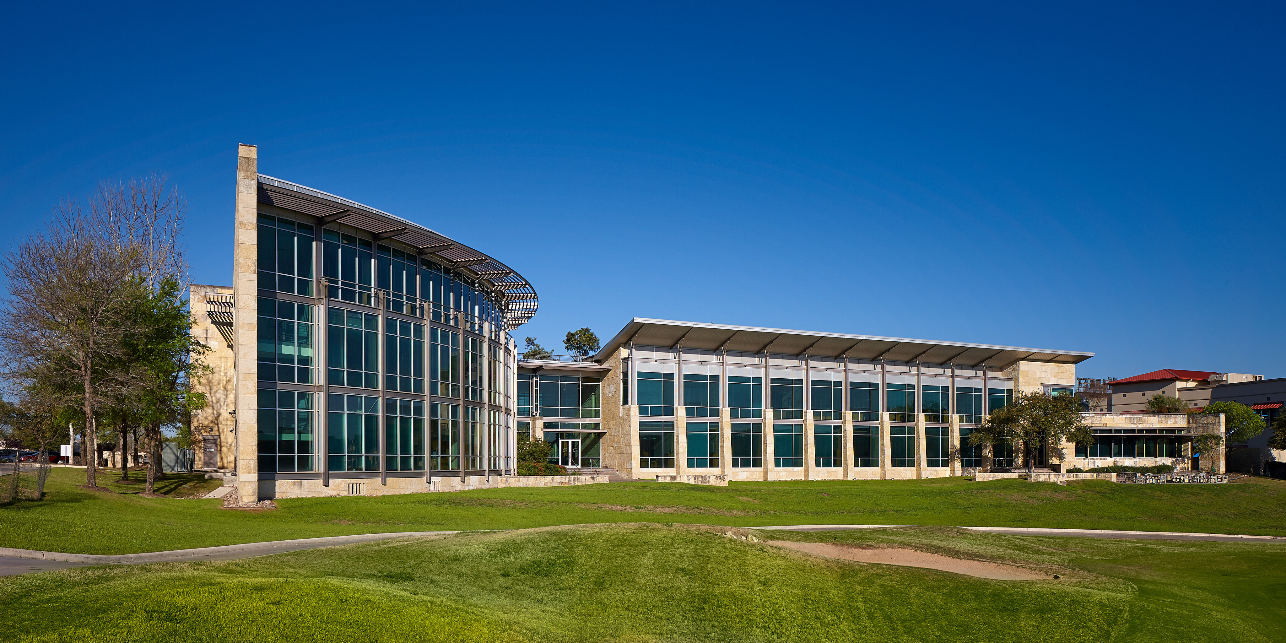 Modern, multi-story building with large glass windows and a curved facade, set against a clear blue sky. The Clear Channel headquarters is surrounded by a well-manicured grassy area and trees.