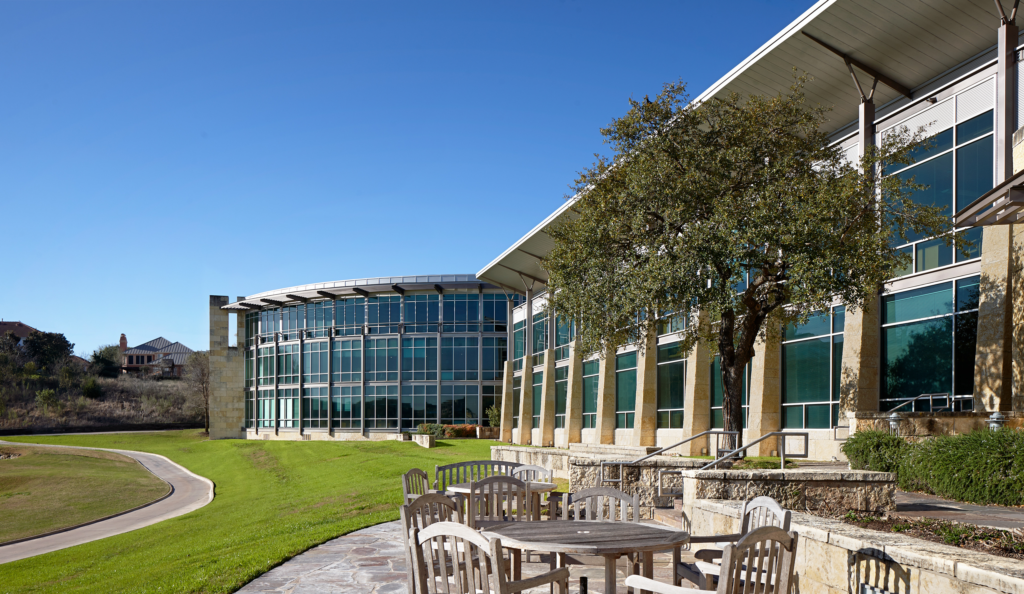 Outdoor patio with wooden tables and chairs overlooking a pathway and the Clear Channel headquarters, featuring large windows and a tree in front. Clear blue sky in the background.