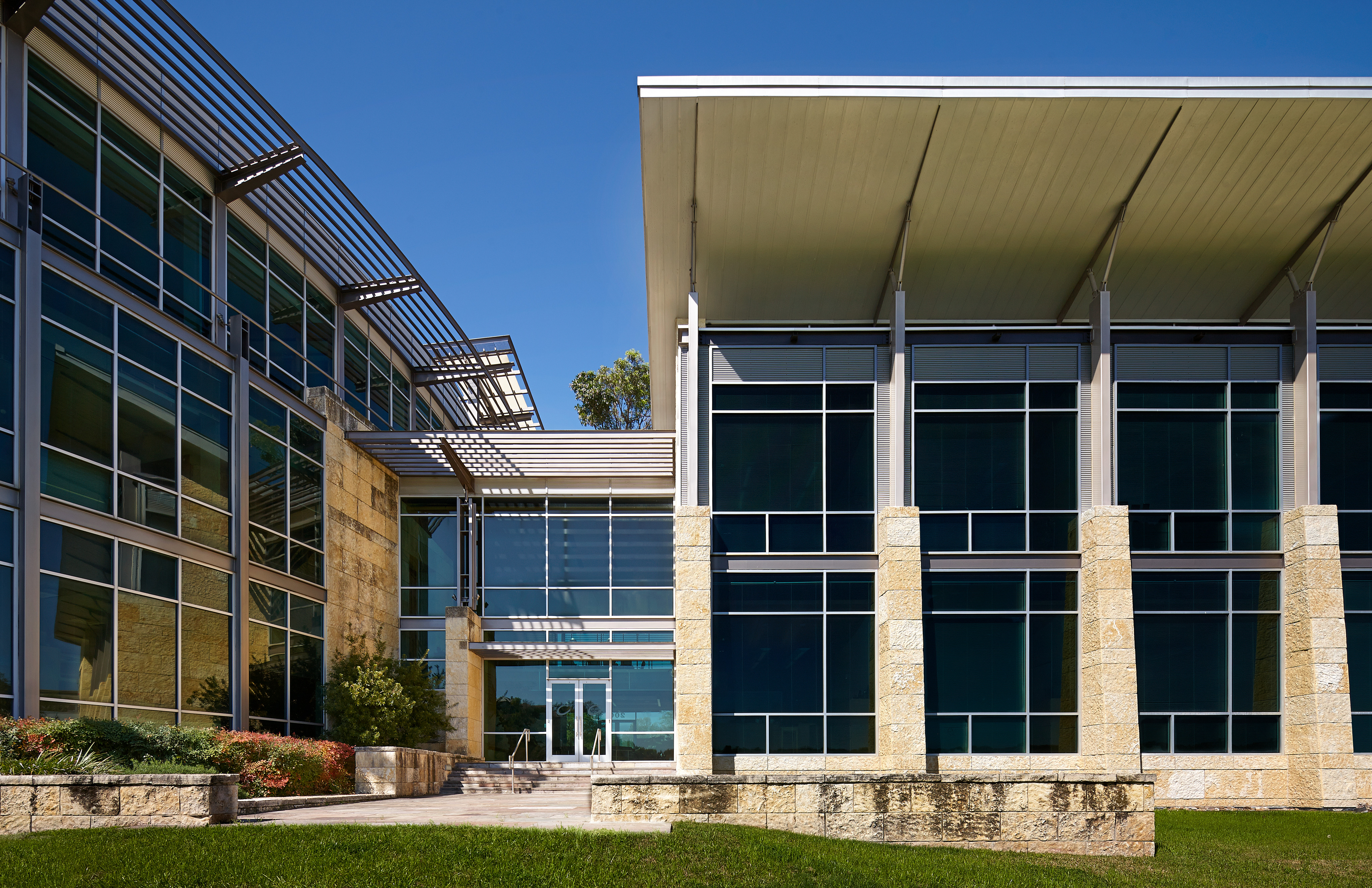 Modern office building with large windows and metal awnings, featuring a mix of stone and glass facade, set against a clear blue sky, serving as the headquarters for a prestigious company.