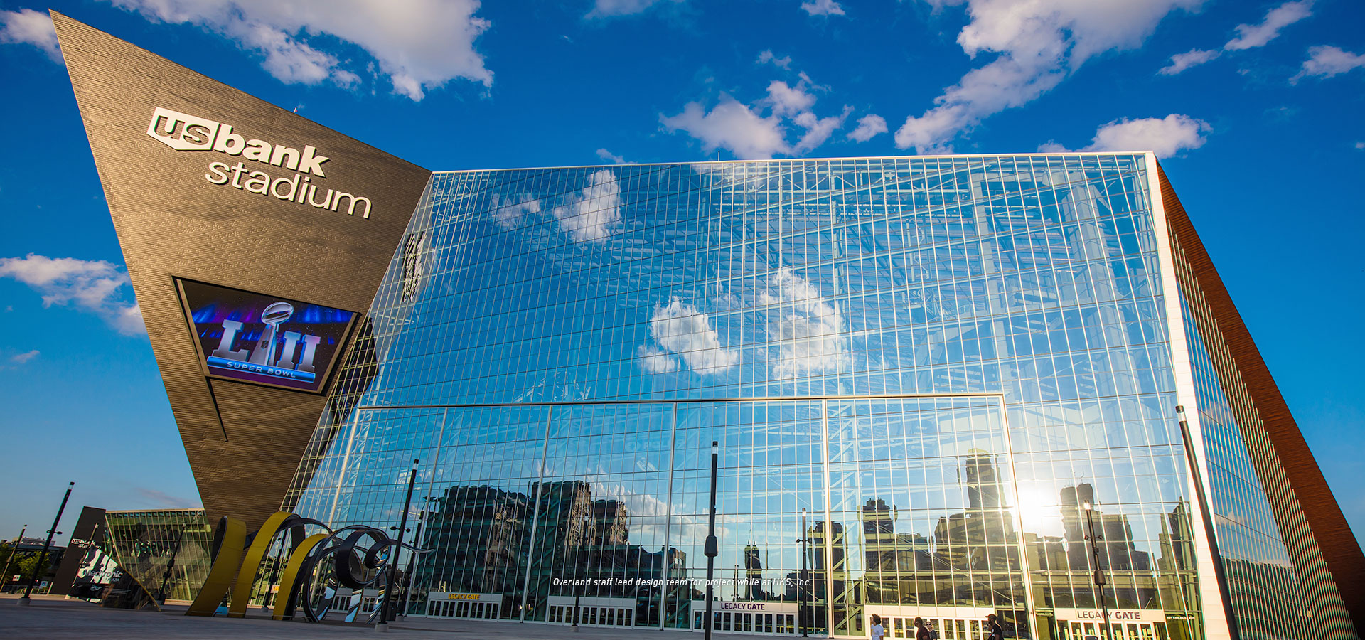 Exterior view of U.S. Bank Stadium with its glass facade reflecting the sky and clouds, under a blue sky with scattered clouds. The iconic US Bank Stadium stands majestically, showcasing its architectural brilliance against the serene backdrop.