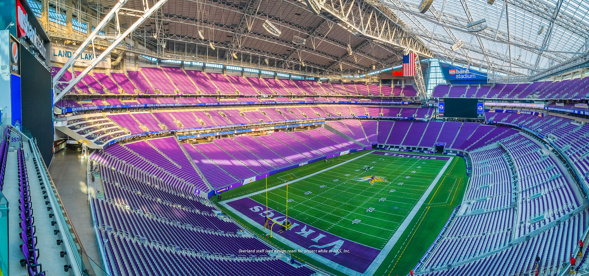 Panoramic view of an empty US Bank Stadium with purple seats, green field, and a large screen on the left. The upper section showcases the retractable roof and structural beams.